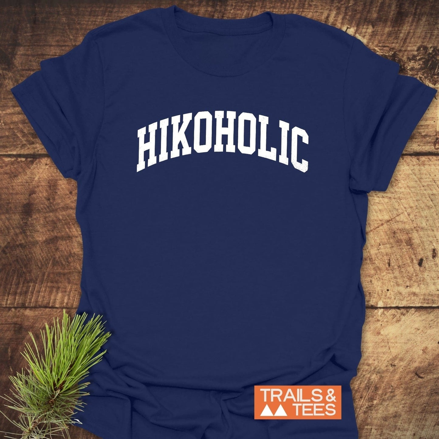 Navy-blue Hikoholic T-shirt with white collegiate lettering, placed on wood with pine needles, exemplifies outdoor adventure spirit.