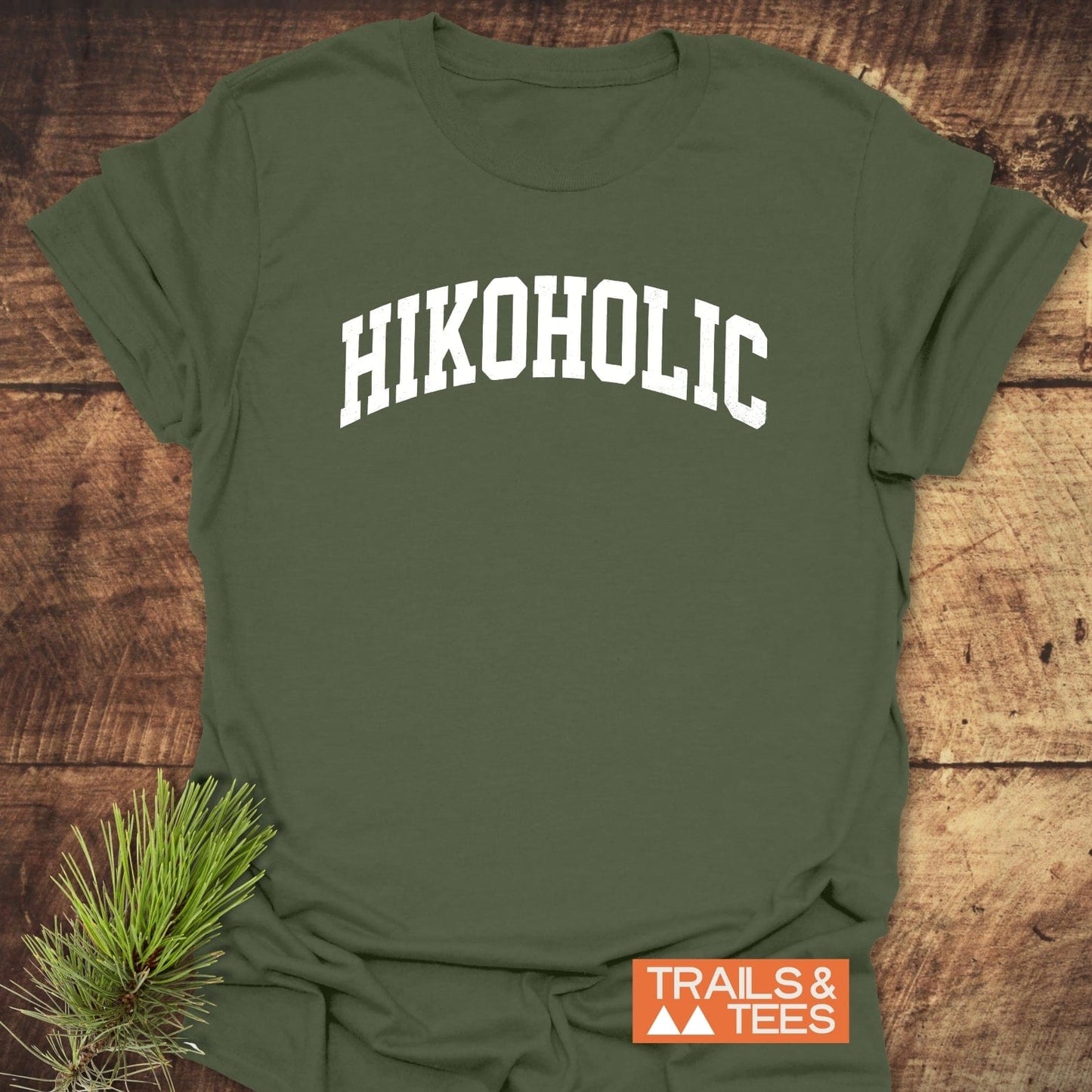 Hikoholic T-Shirt on wooden surface, featuring white collegiate lettering and orange Trails & Tees logo, styled with a pine sprig for an outdoor theme.