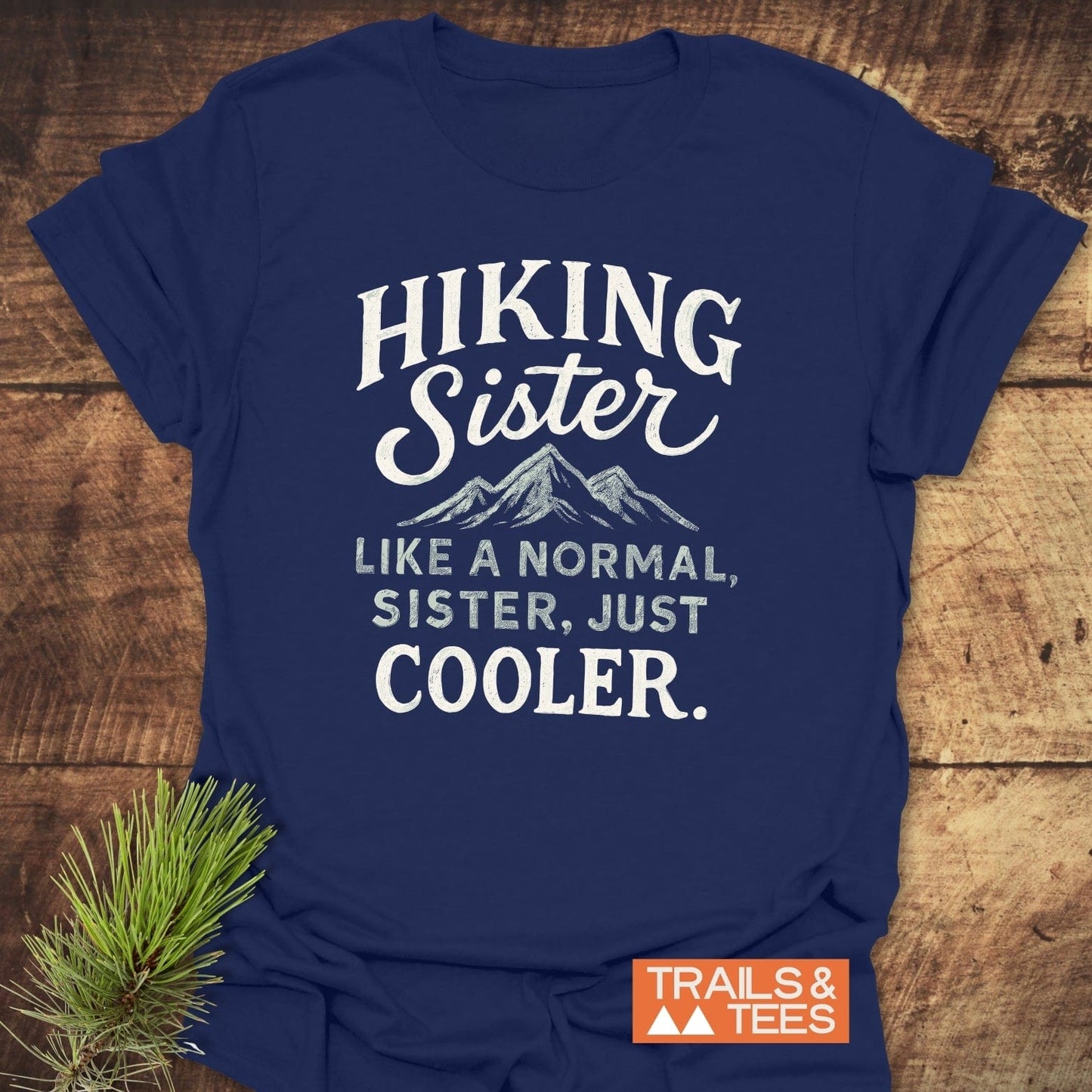 Navy-blue Hiking Sister T-shirt with distressed mountain graphic and slogan, laid flat on wood with pine twig; branded TRAILS & TEES in corner.