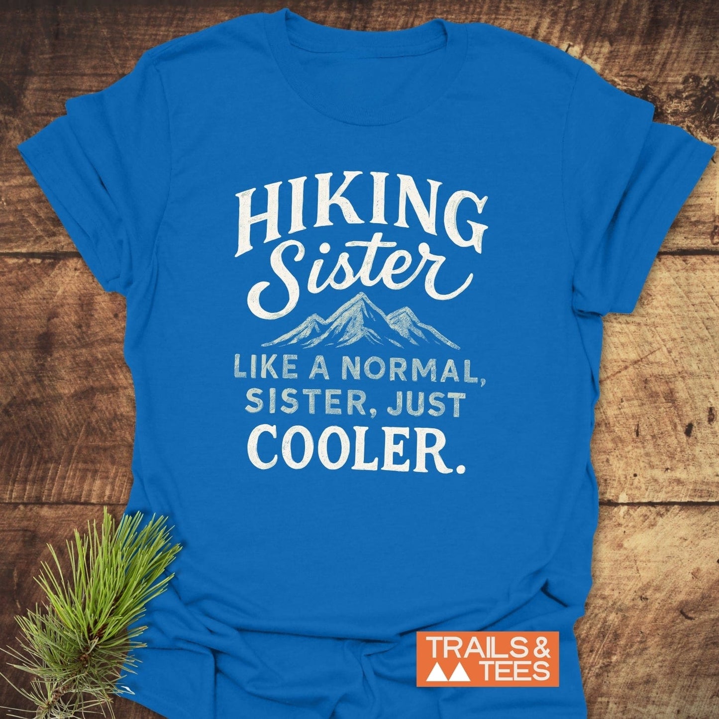 Hiking Sister T-Shirt on a wooden surface with a mountain graphic and text. Features a small Trails & Tees logo and a pine needle accent.