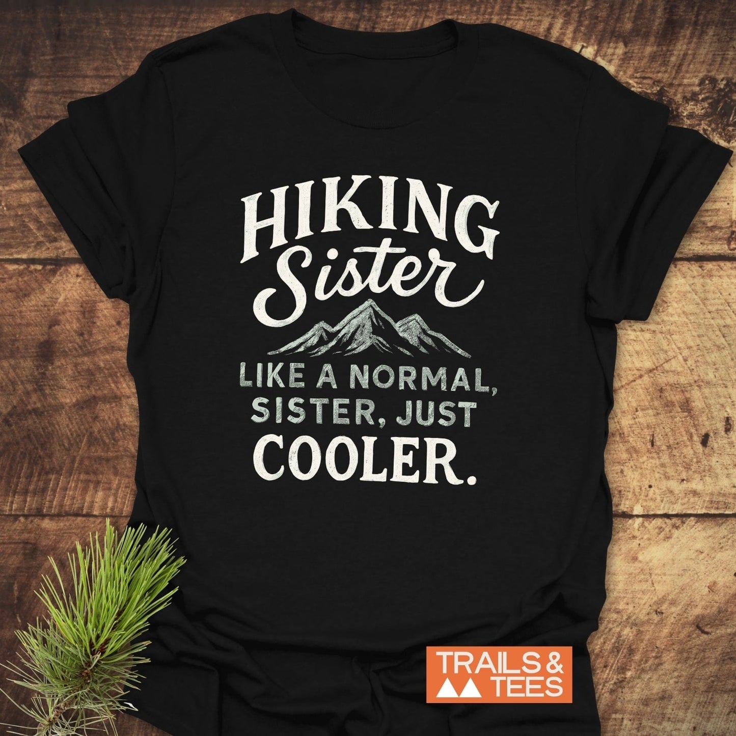 Hiking Sister T-Shirt with mountain graphic, laid flat. Features soft ring-spun cotton and durable print for outdoor enthusiasts. TRAILS & TEES logo in corner.
