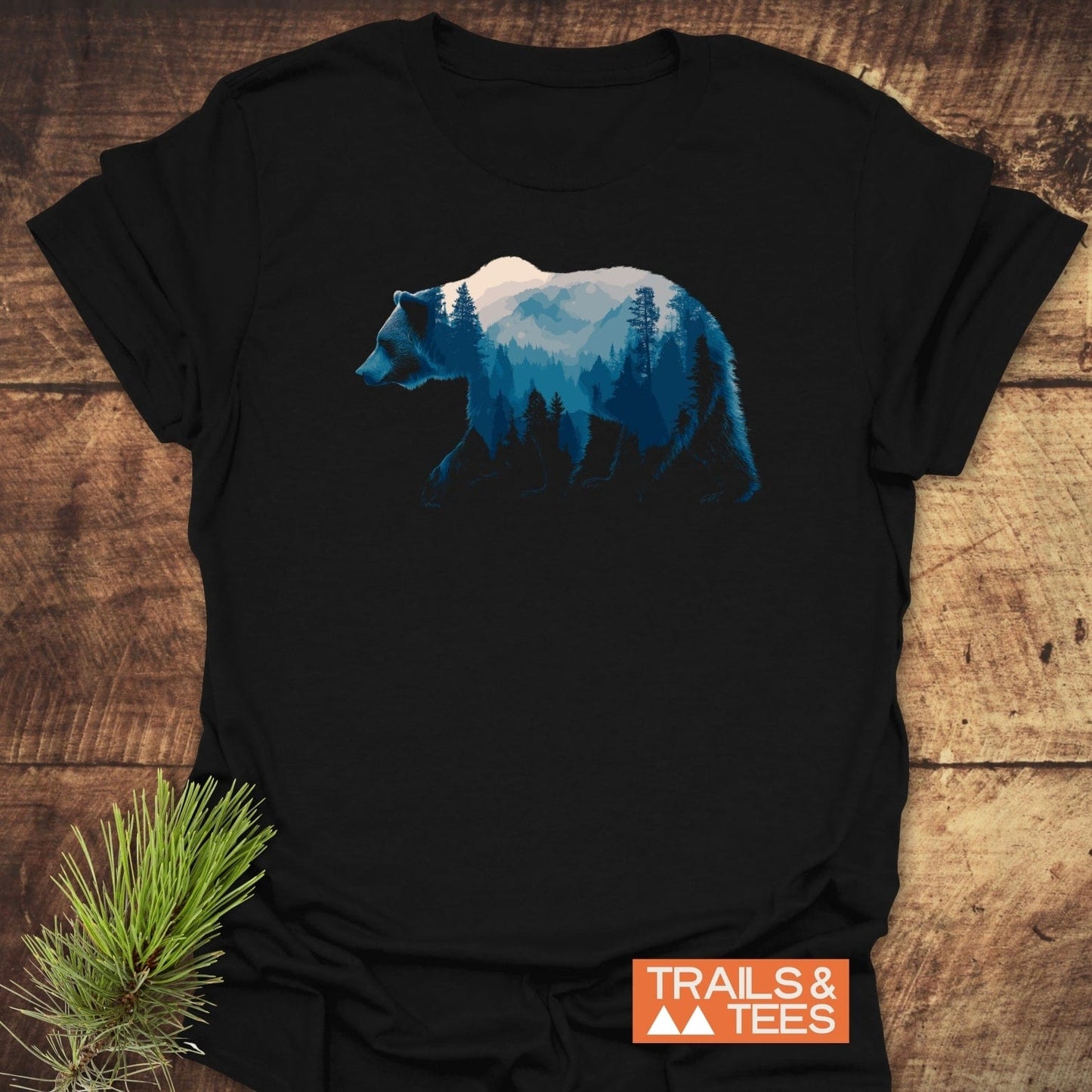 Grizzly Silhouette T-Shirt featuring a bear with mountain scene graphic, laid on wood. Includes TRAILS & TEES badge. Made from soft, lightweight cotton.