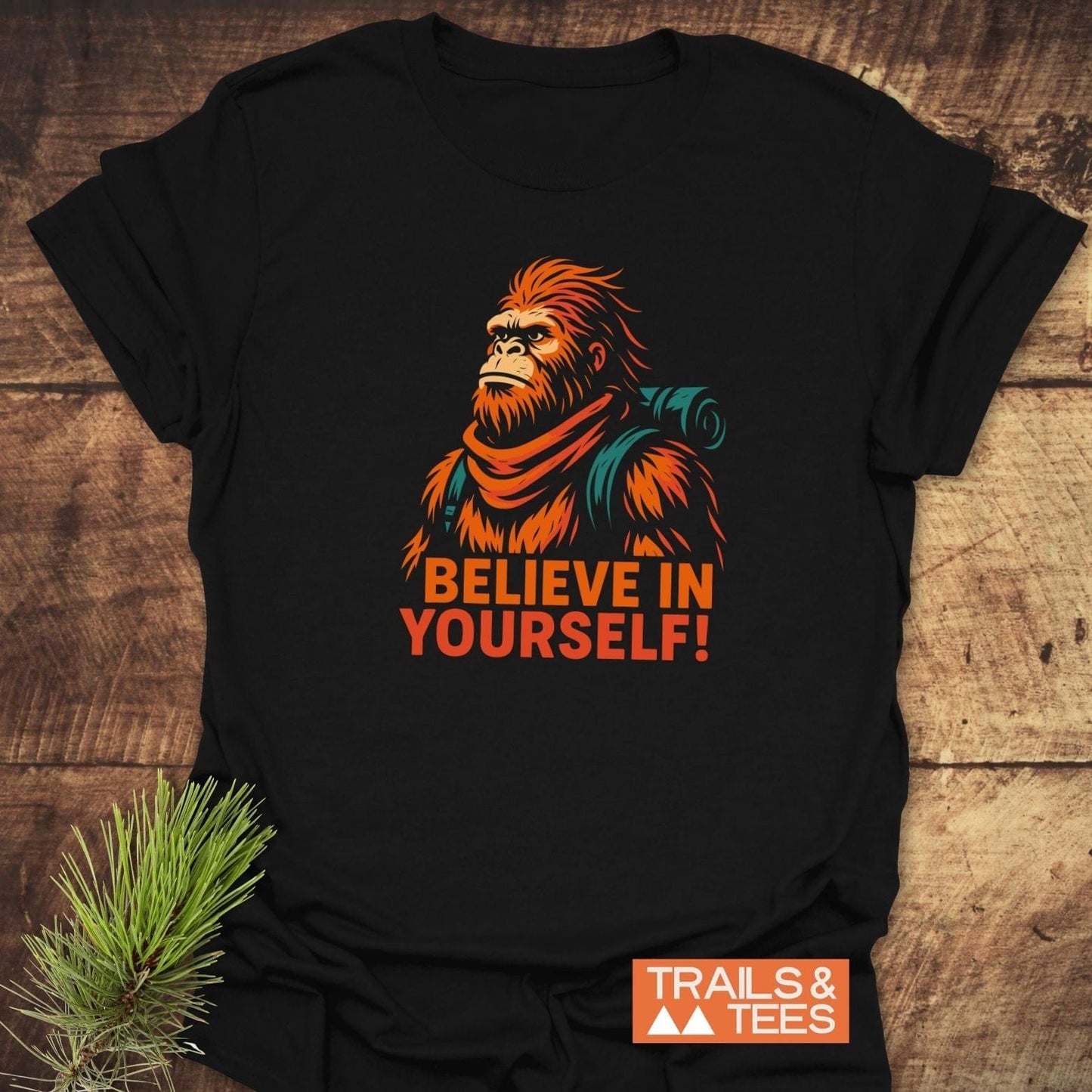 Believe In Yourself Bigfoot T-Shirt with a stylized Sasquatch illustration, crafted from soft cotton for comfort and durability, laid on a rustic wood surface.