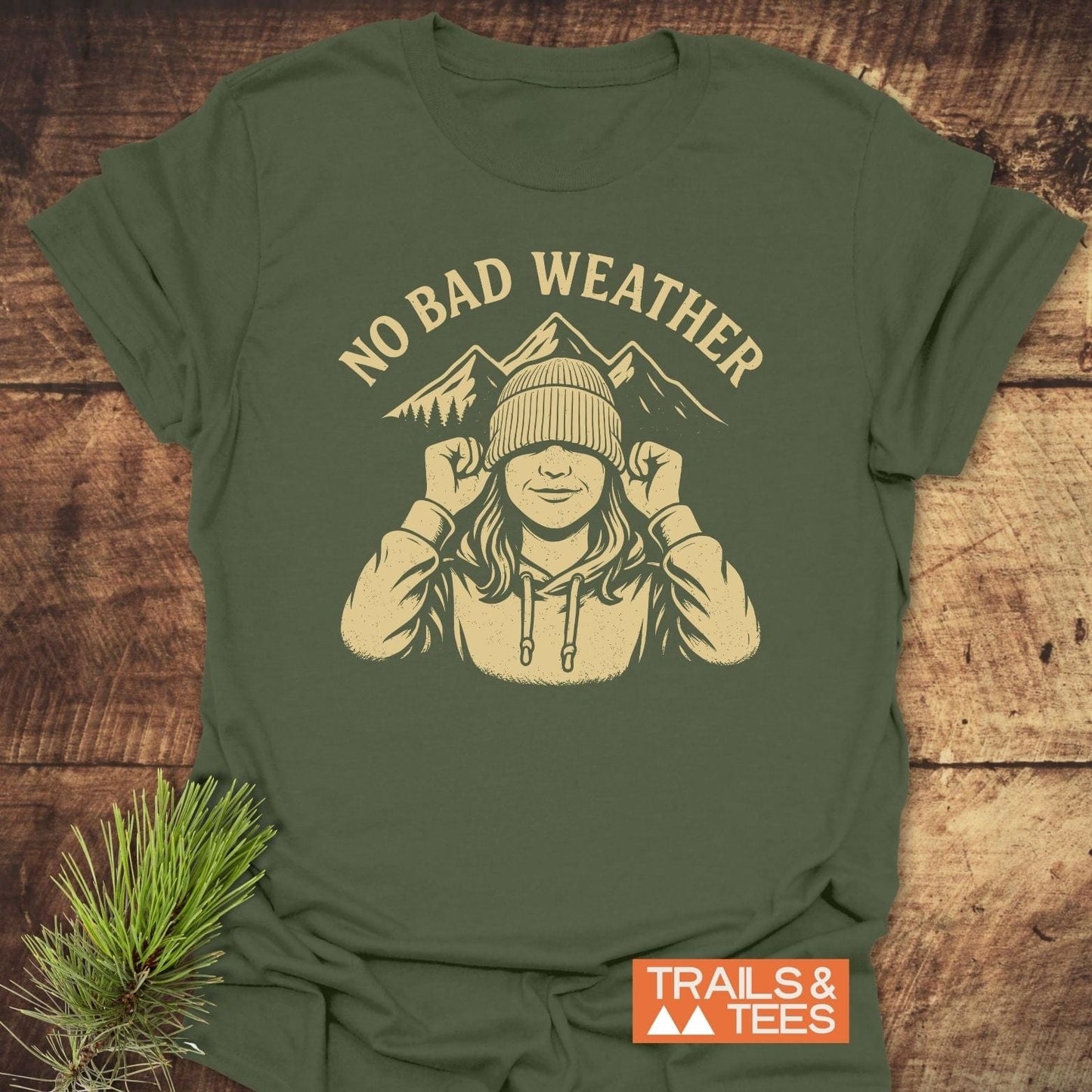 No Bad Weather T-Shirt featuring a cream graphic on green fabric, showcasing a person in a cap with mountains. Includes Trails & Tees logo.