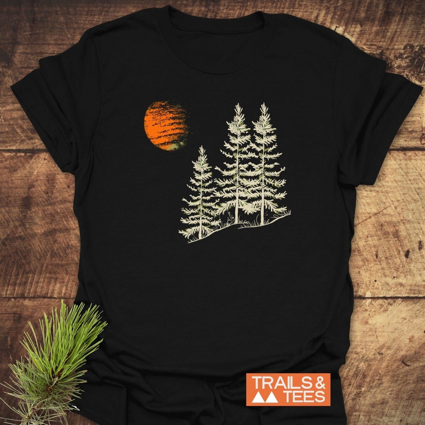 Forest And Moon T-Shirt featuring three pine trees and an orange circle design, crafted from soft cotton, styled with a pine sprig on rustic wood.