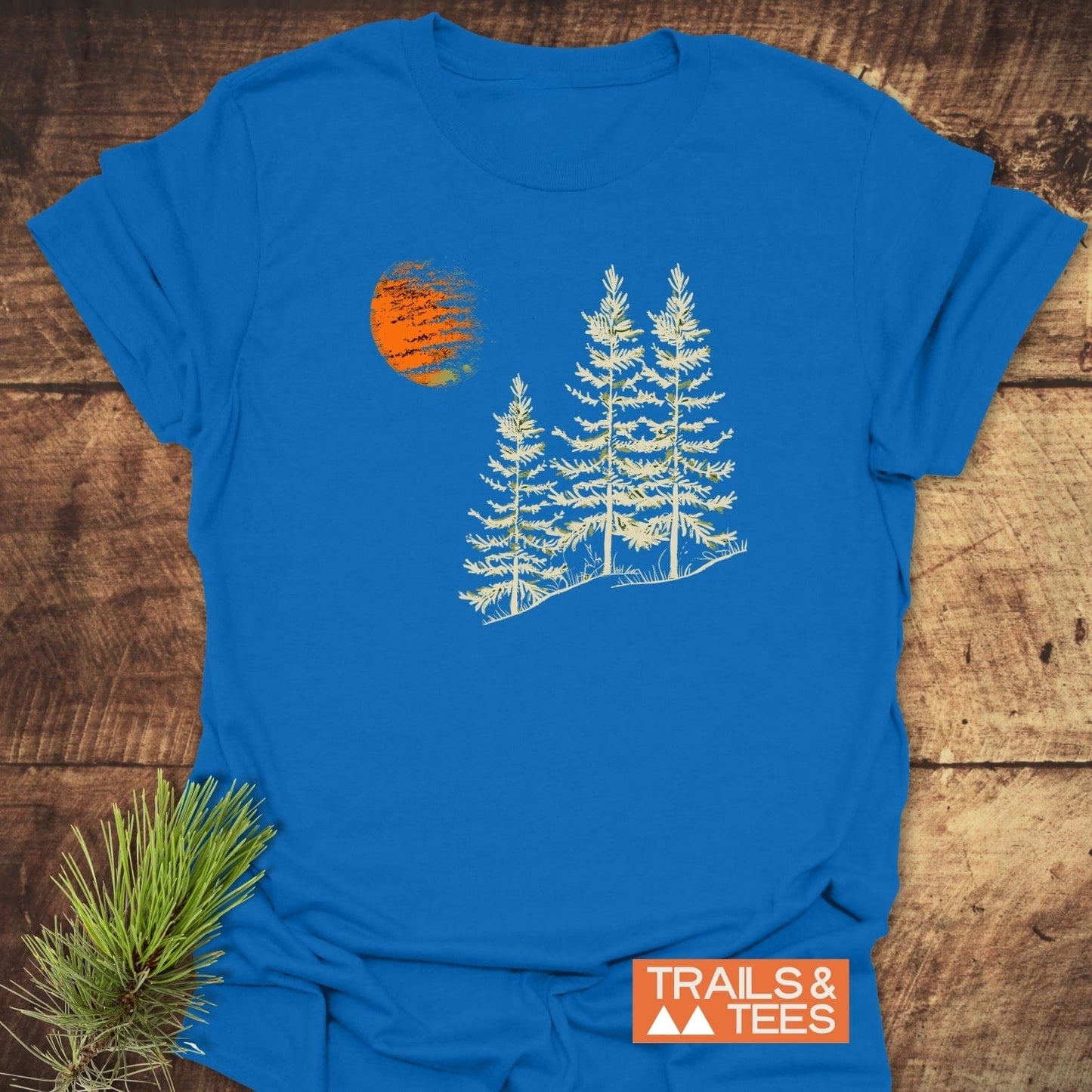 Forest And Moon T-Shirt featuring cream evergreen trees and an orange sun, displayed on a wooden surface, symbolizing outdoor adventure and crafted for comfort.