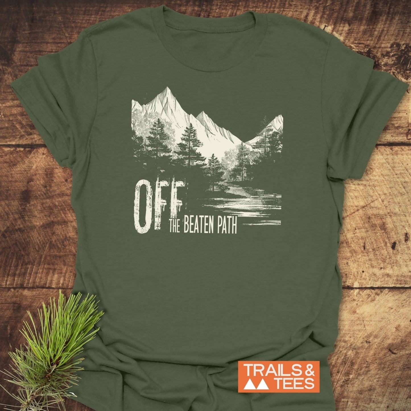 Off The Beaten Path T-Shirt featuring mountain and pine graphic, slogan, and Trails & Tees logo, styled with pine needles on a wooden surface.