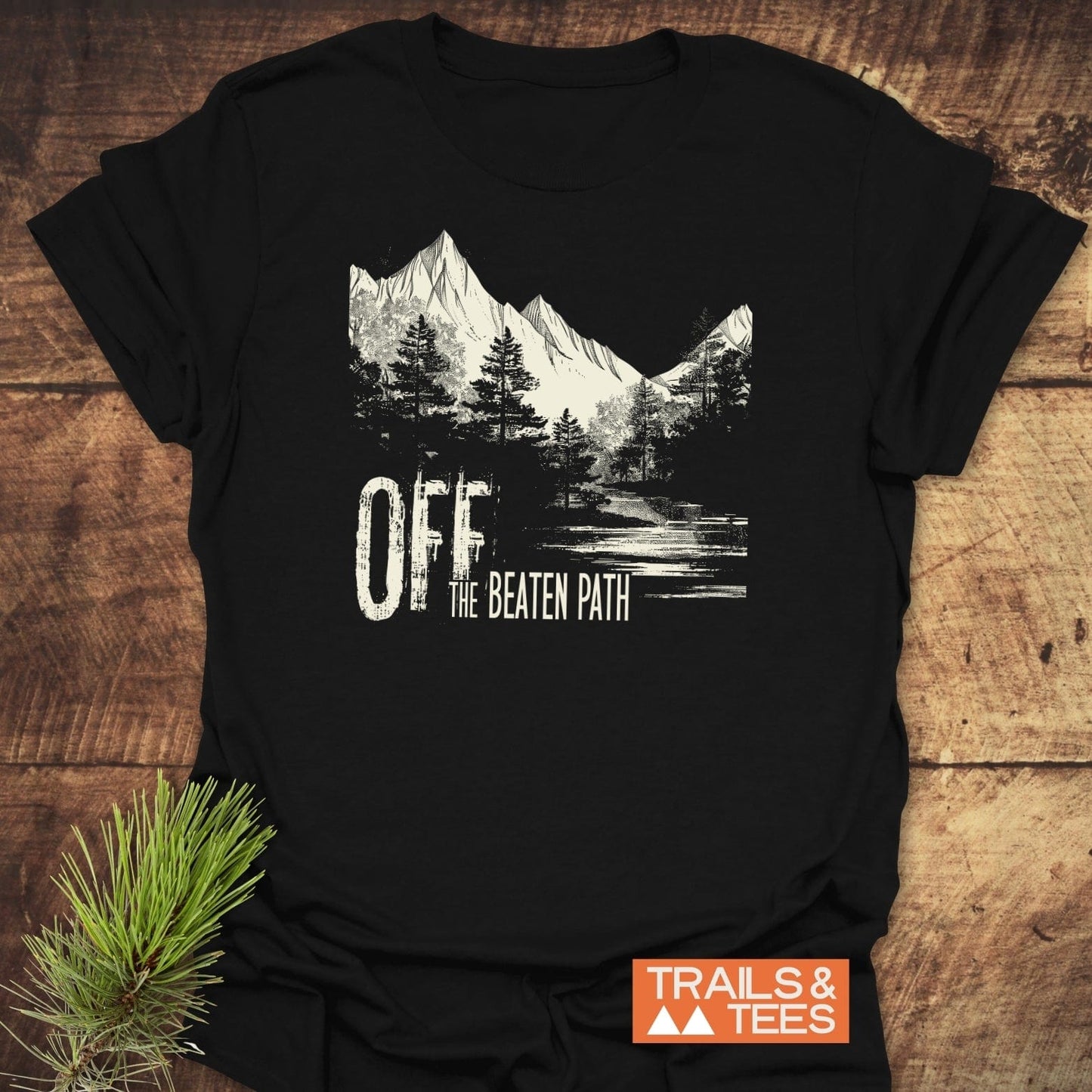 Off The Beaten Path T-Shirt featuring a mountain and forest scene, durable DTF print, and Trails & Tees logo, ideal for outdoor enthusiasts.