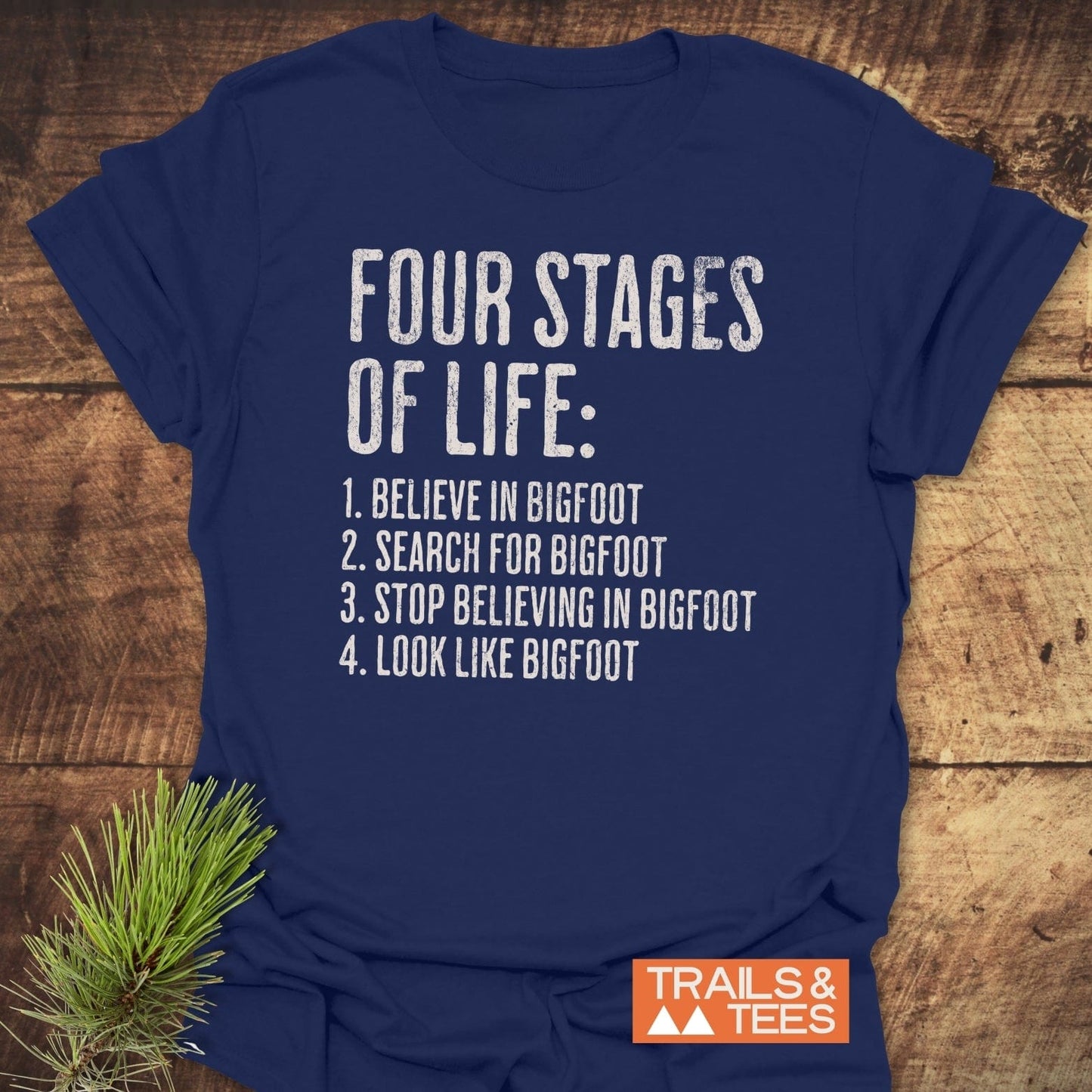 Four Stages Of Life T-Shirt with humorous Bigfoot slogan printed on soft navy-blue cotton, featuring Trails & Tees logo and pine sprig decoration.