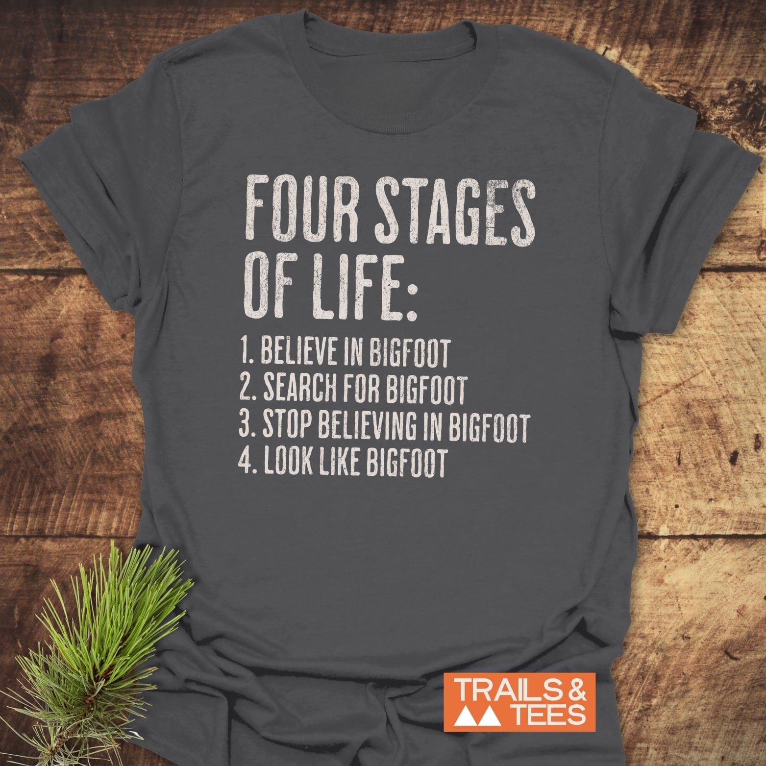 Four Stages Of Life T-Shirt on wood, featuring distressed text about Bigfoot. Includes Trails & Tees logo. Pine sprig accentuates outdoor theme.