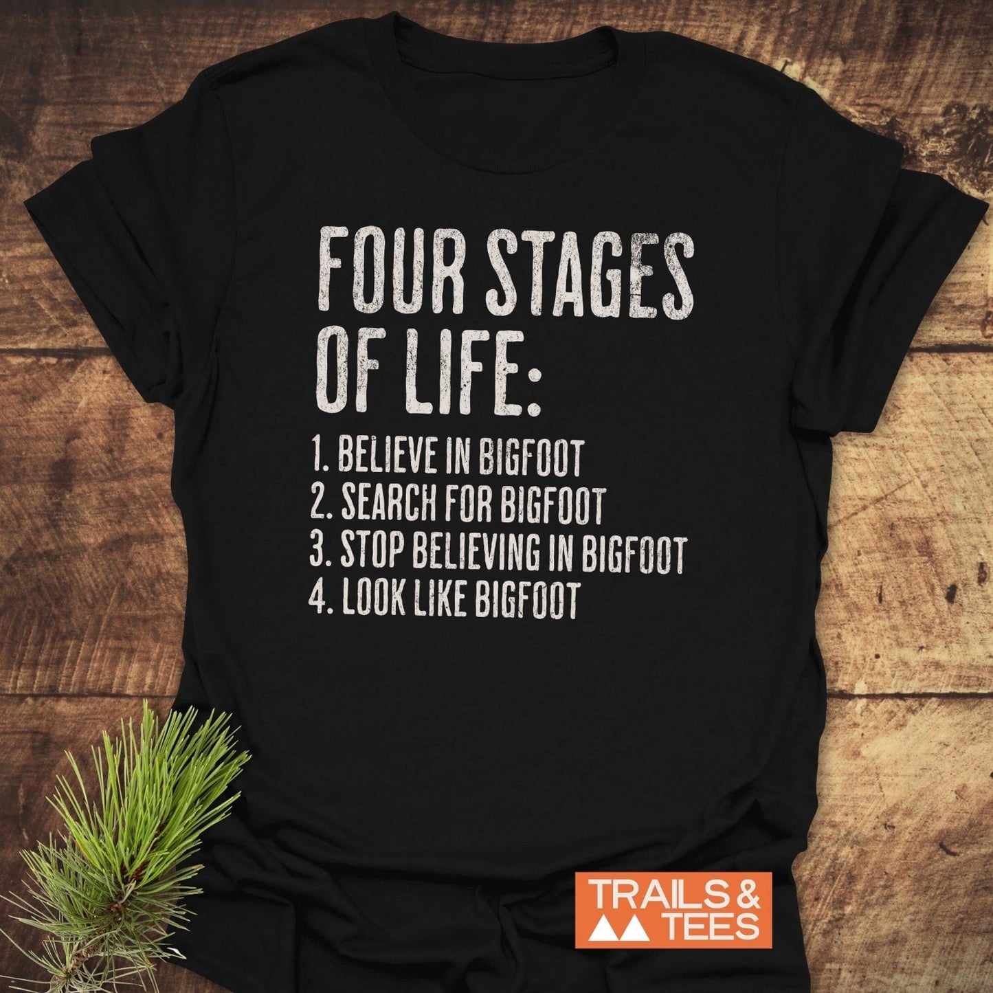 Four Stages Of Life T-Shirt with humorous Bigfoot-themed text, featuring durable print and Trails & Tees logo, designed for comfort and outdoor adventures.