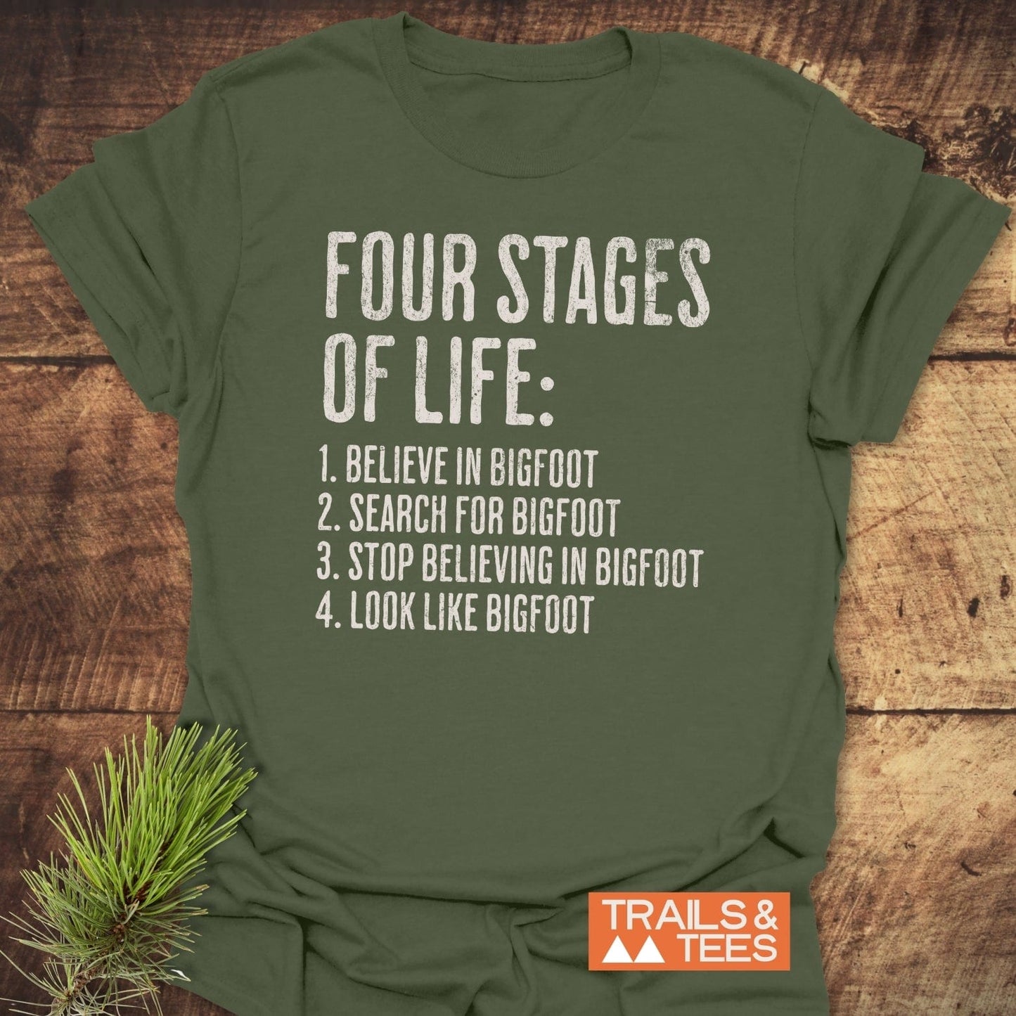 Four Stages Of Life T-Shirt featuring distressed white lettering, orange Trails & Tees logo, crafted from soft cotton for comfort and durability, styled with pine sprigs.