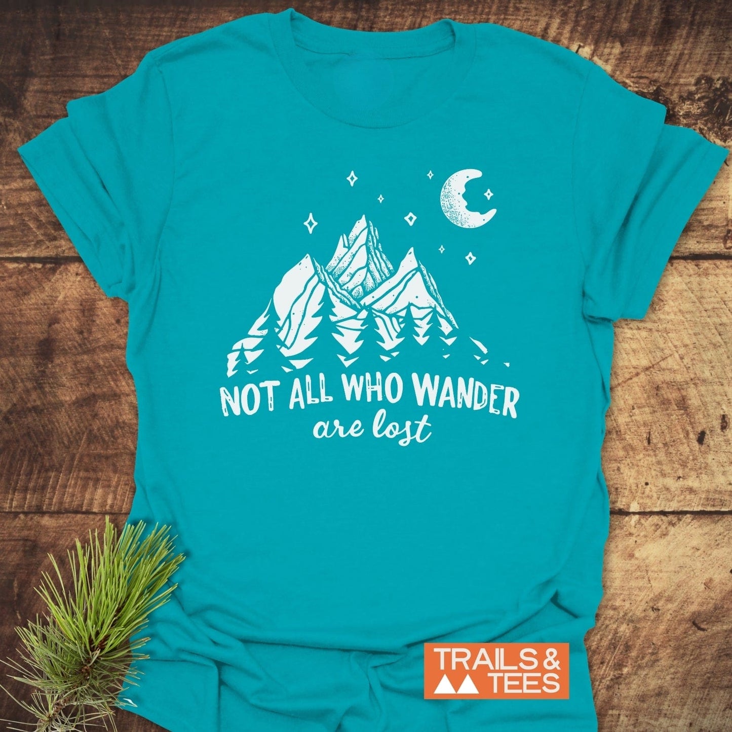 Not All Who... Nightsky T-Shirt with a mountain design and NOT ALL WHO WANDER are lost print on turquoise-blue cotton, laid on rustic wood.