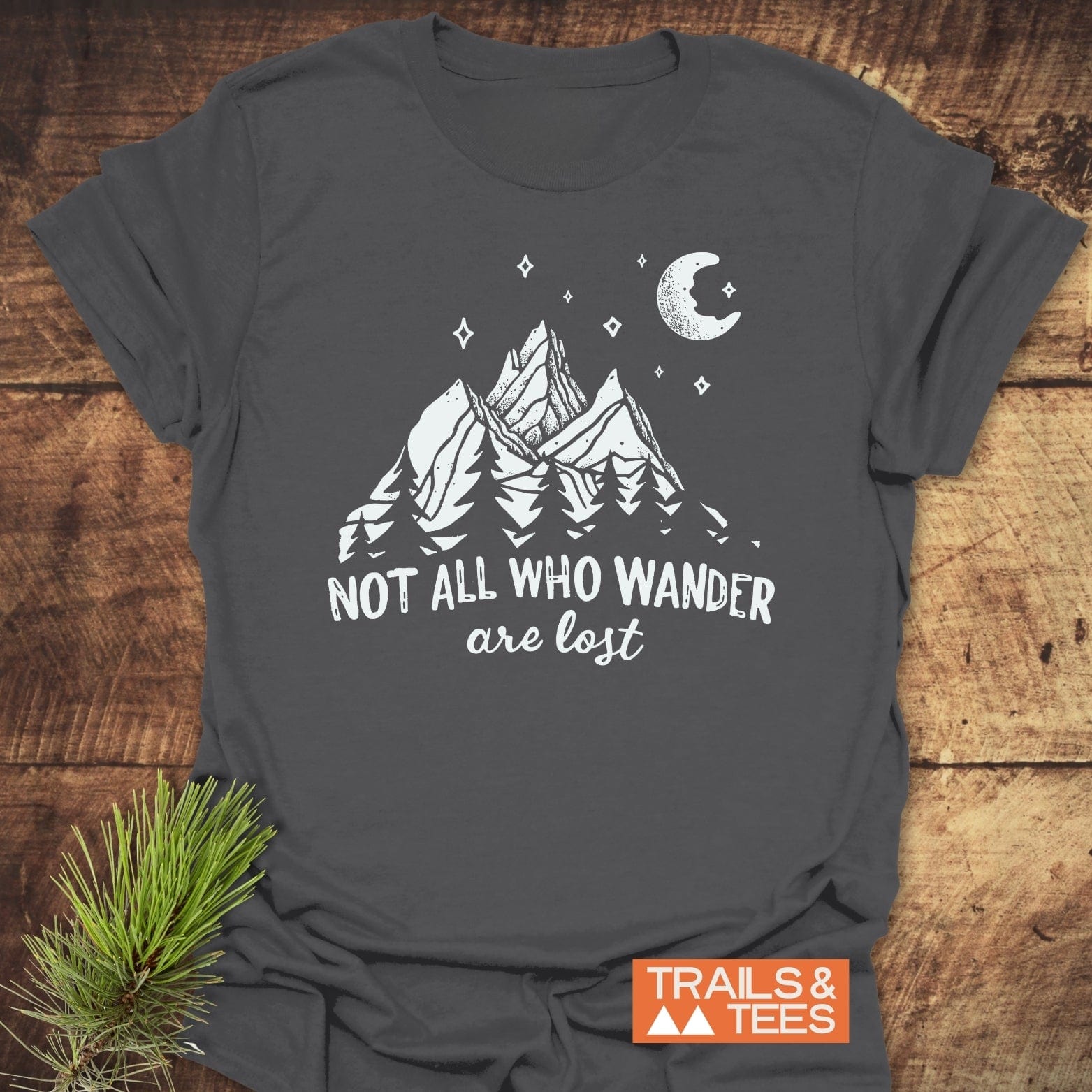 Not All Who Wander Nightsky T-Shirt with mountain and pine design; soft, ring-spun cotton, pre-shrunk, durable print, laid on wooden surface with pine sprig.