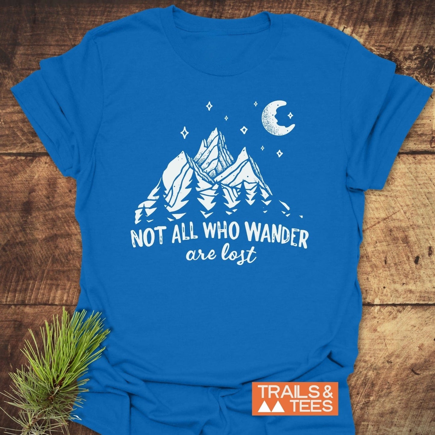 Not All Who... Nightsky T-Shirt on wood, featuring a mountain scene and slogan. Includes Trails & Tees label and pine sprig, emphasizing outdoor adventure.