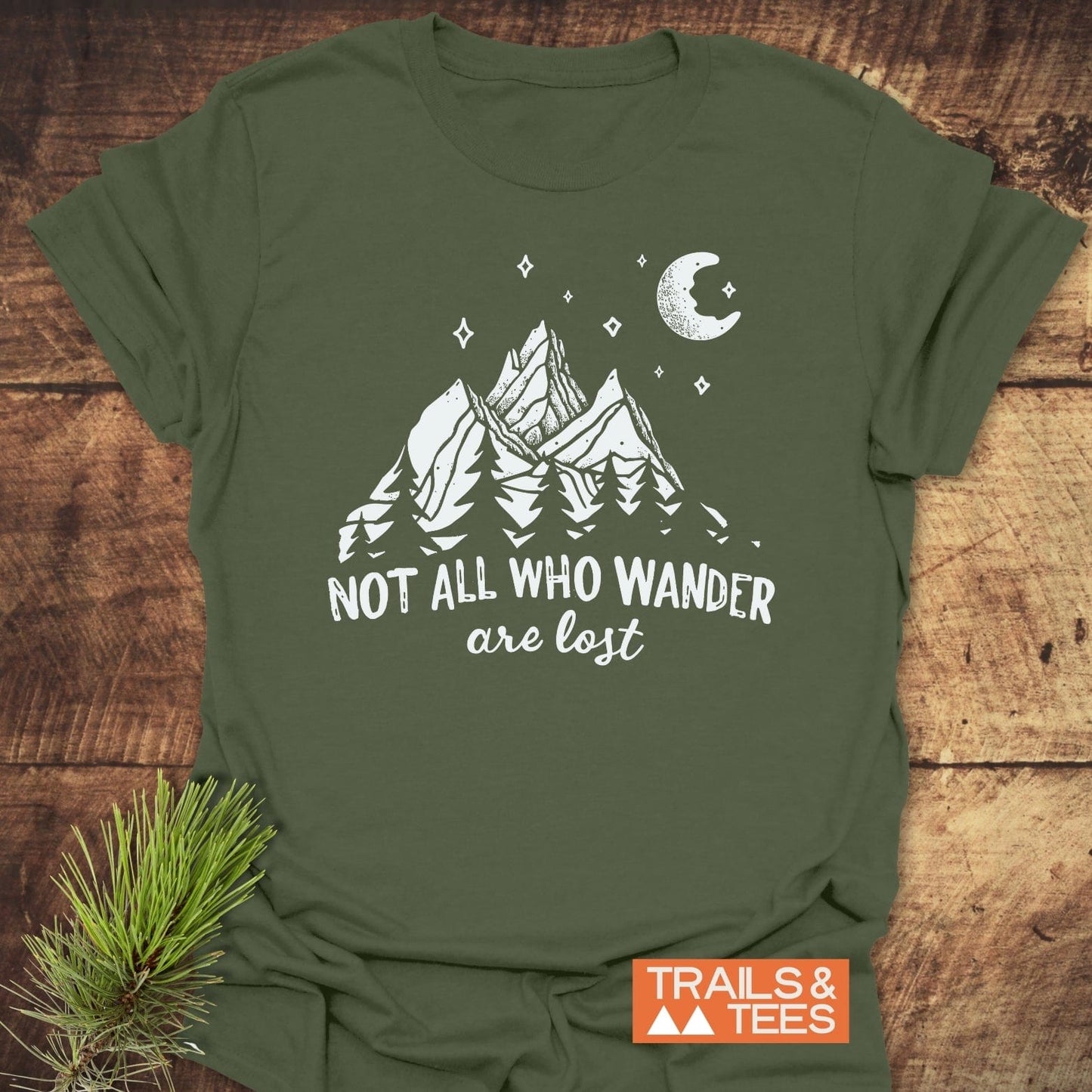Army-green T-shirt with white graphic of mountains, trees, stars, and moon; reads “Not all who wander are lost.” Featuring comfort-focused, durable design by TRAILS & TEES.