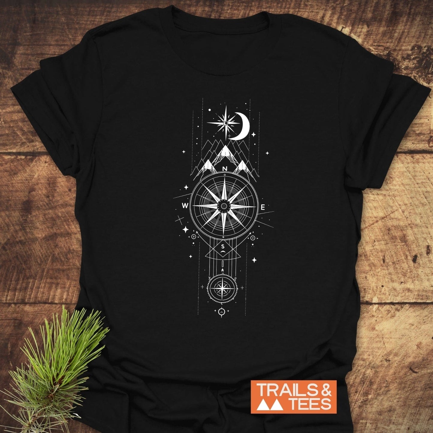 Celestial Compass T-Shirt featuring a white compass and mountain design on a black tee, laid on wood with a pine sprig and TRAILS & TEES logo.