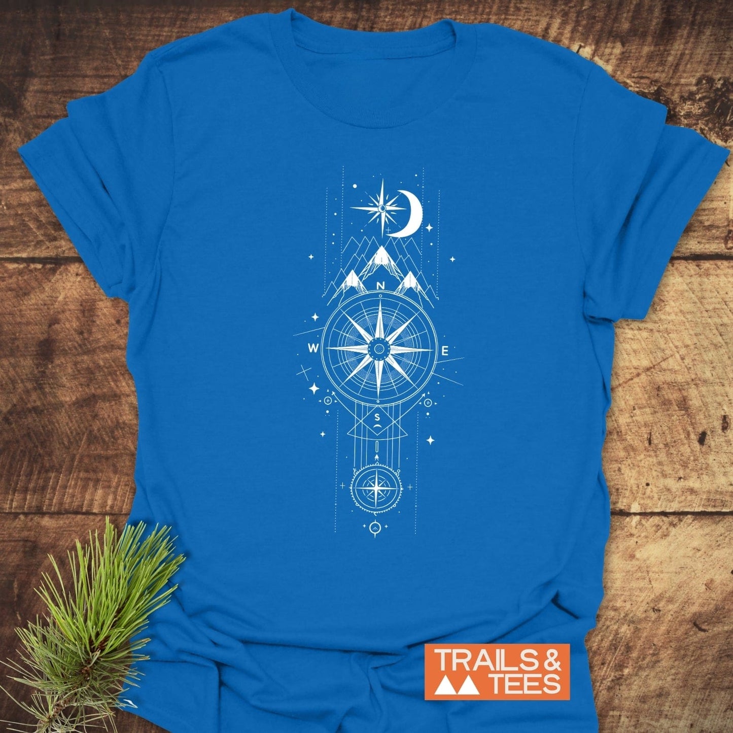 Celestial Compass T-Shirt featuring a white compass design, mountain peaks, and celestial motifs, laid on wood, embodying adventure-ready, soft ring-spun cotton for outdoor enthusiasts.