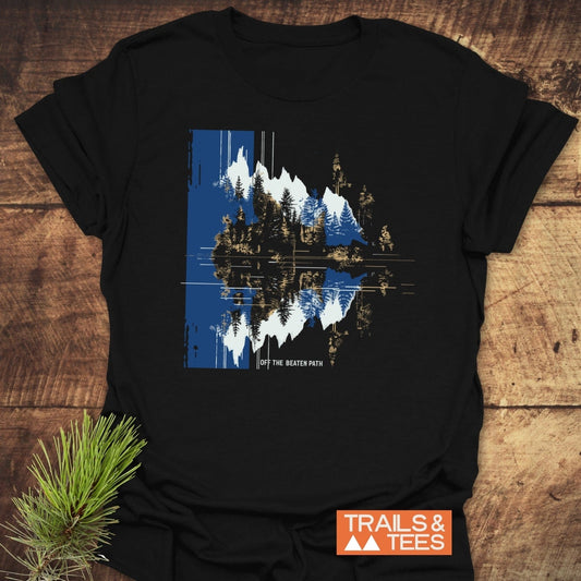 Minimalist Mountain Reflection T-Shirt with a stylized mountain and forest design, slogan “OFF THE BEATEN PATH,” and TRAILS & TEES logo on wooden surface.