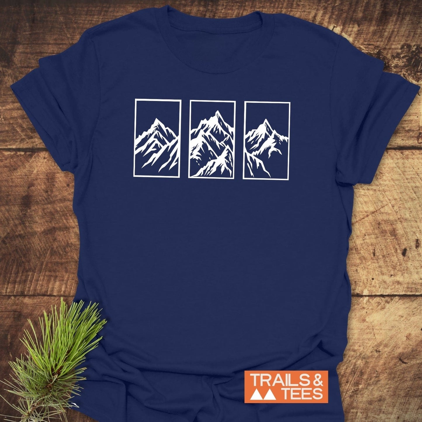 Navy-blue Mountain Triptych T-Shirt with white mountain illustrations, orange TRAILS & TEES logo, laid on wood. Features durable, pre-shrunk cotton for comfort.