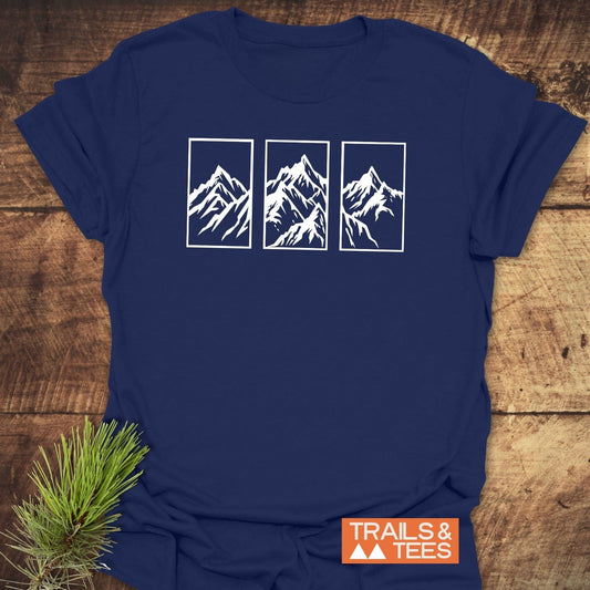 Navy-blue Mountain Triptych T-Shirt with white mountain illustrations, orange TRAILS & TEES logo, laid on wood. Features durable, pre-shrunk cotton for comfort.