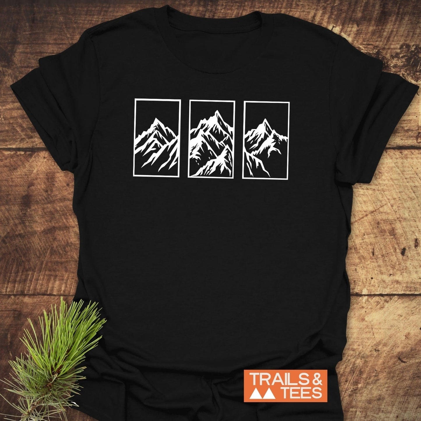 Mountain Triptych T-Shirt featuring stylized mountain peaks in white frames, laid flat with a pine twig. Crafted for outdoor adventures, showcasing Trail-tested comfort and durability.
