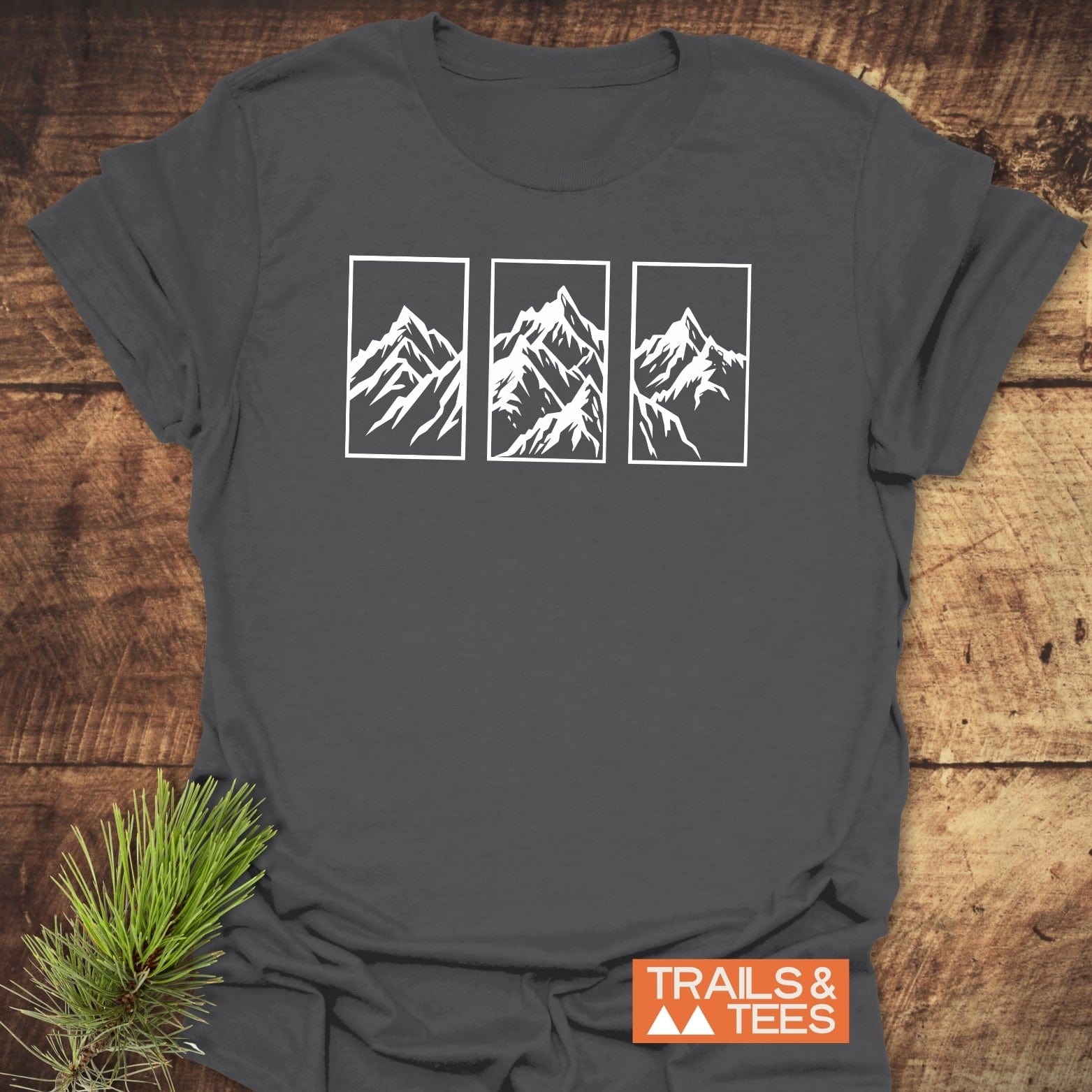 Mountain Triptych T-Shirt on wood surface, features stylized mountain peaks on chest, with TRAILS & TEES logo and pine sprig accenting the display.