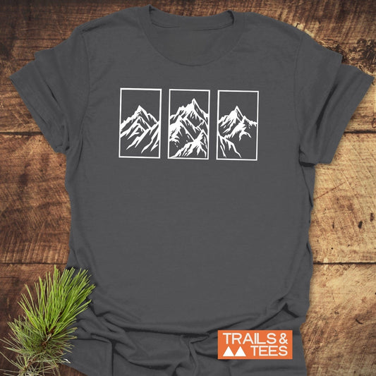 Mountain Triptych T-Shirt on wood surface, features stylized mountain peaks on chest, with TRAILS & TEES logo and pine sprig accenting the display.