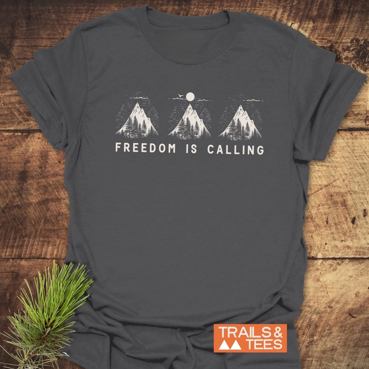 Freedom Is Calling T-Shirt with mountain peaks graphic, laid on wood. Crafted from soft, lightweight cotton, pre-shrunk for fit, featuring durable prints.