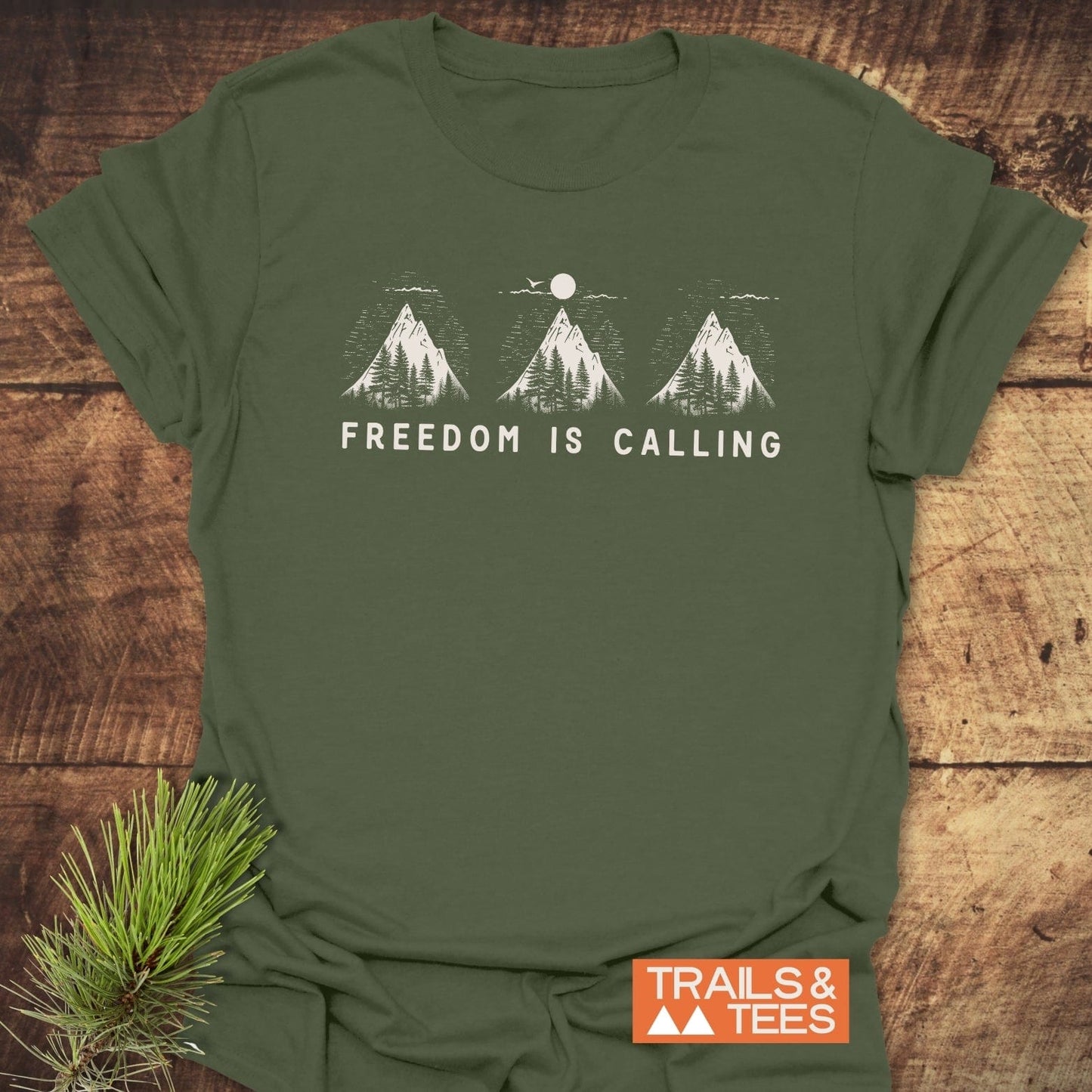 Freedom Is Calling T-Shirt featuring mountain silhouettes and slogan, laid flat with a pine sprig; crafted from soft ring-spun cotton for outdoor enthusiasts.