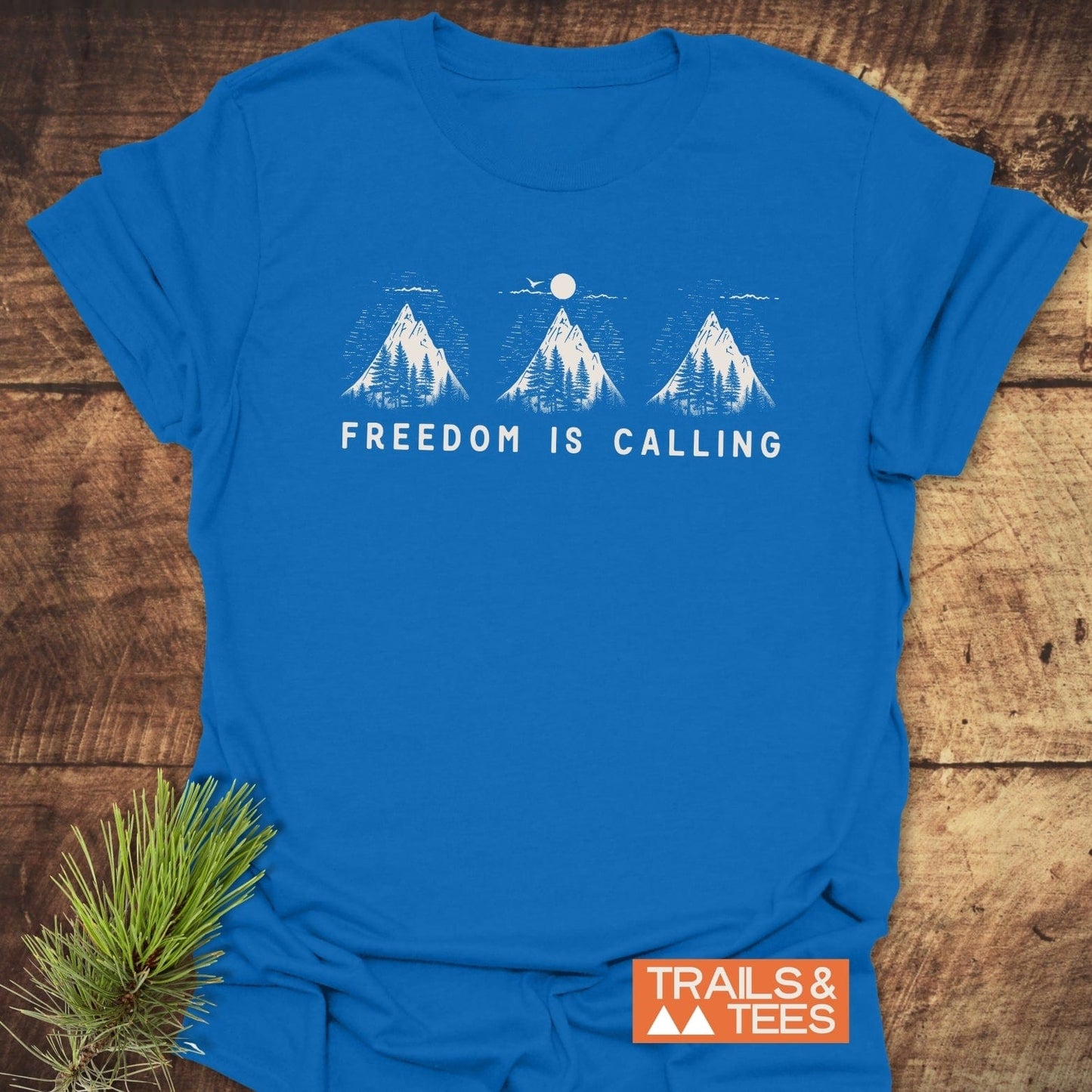 Freedom Is Calling T-Shirt on rustic wood, featuring a white mountain graphic with trees and sun, and TRAILS & TEES logo, embodying outdoor spirit and adventure.