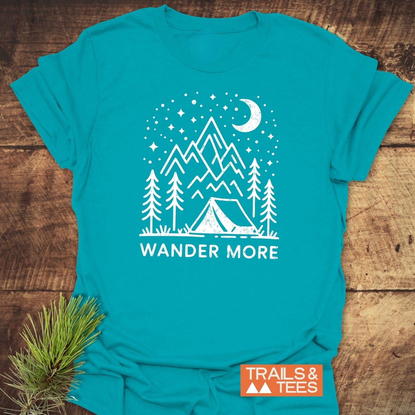 Wander More T-Shirt with crescent moon, stars, mountains, and trees graphic; slogan “WANDER MORE”; “TRAILS & TEES” logo visible; pine sprig adjacent.