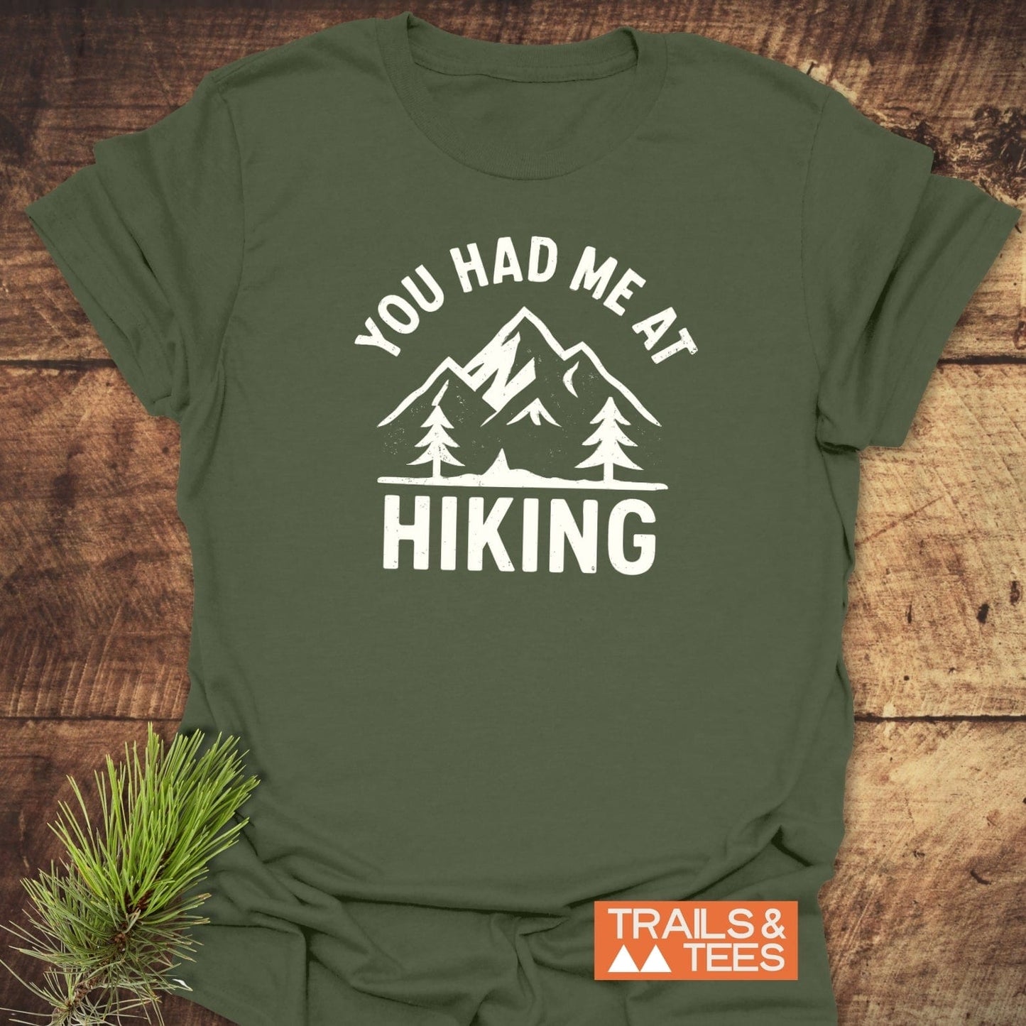 You Had Me At Hiking T-Shirt flat-lay, featuring mountain-and-tree graphic. Includes Trails & Tees logo and pine sprig on rustic wood surface.
