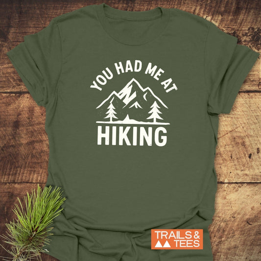 You Had Me At Hiking T-Shirt flat-lay, featuring mountain-and-tree graphic. Includes Trails & Tees logo and pine sprig on rustic wood surface.