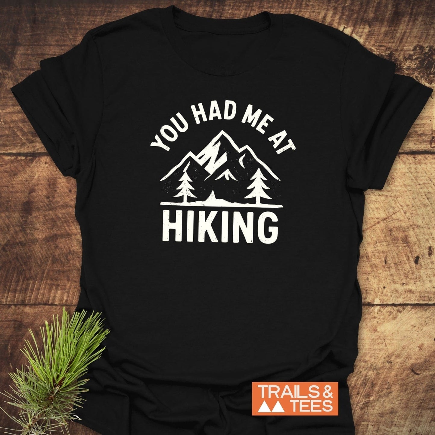 You Had Me At Hiking T-Shirt featuring a mountain-and-pine-tree graphic and slogan, laid flat with a rustic pine sprig accent for an outdoorsy feel.