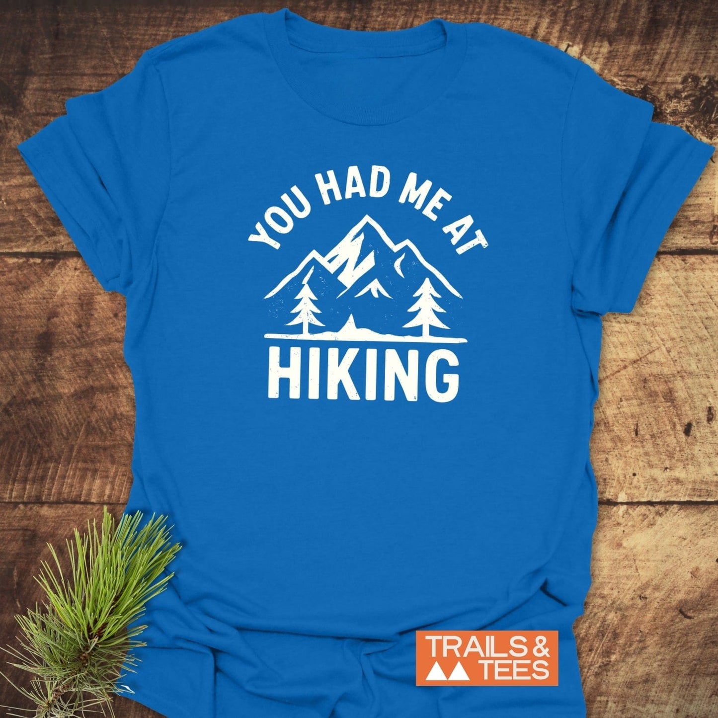 You Had Me At Hiking T-Shirt featuring a mountain range and pine trees design, emphasizing outdoor adventure. Displayed with Trails & Tees logo and spruce garnish.