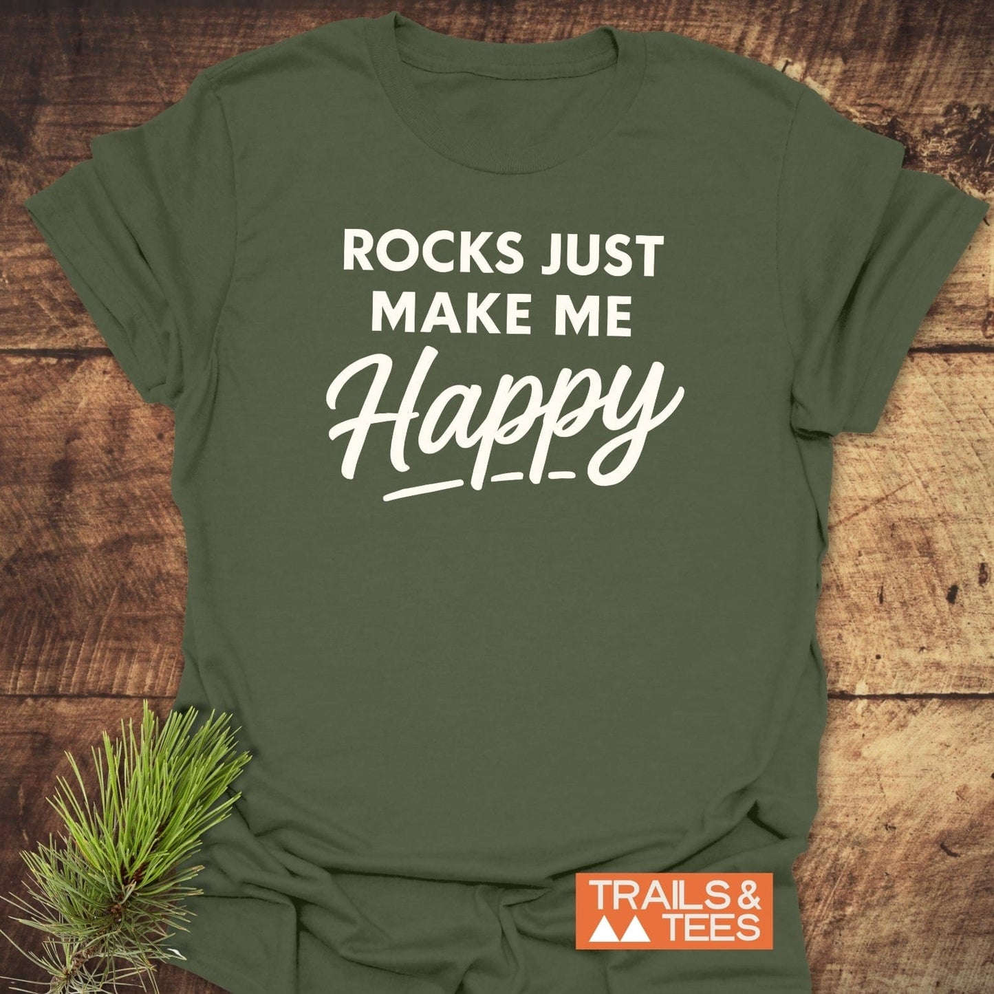 Rocks Make Me Happy T-Shirt with bold white lettering on olive-green fabric, laid on wood. Features durable prints, lightweight cotton, promoting outdoor adventure.