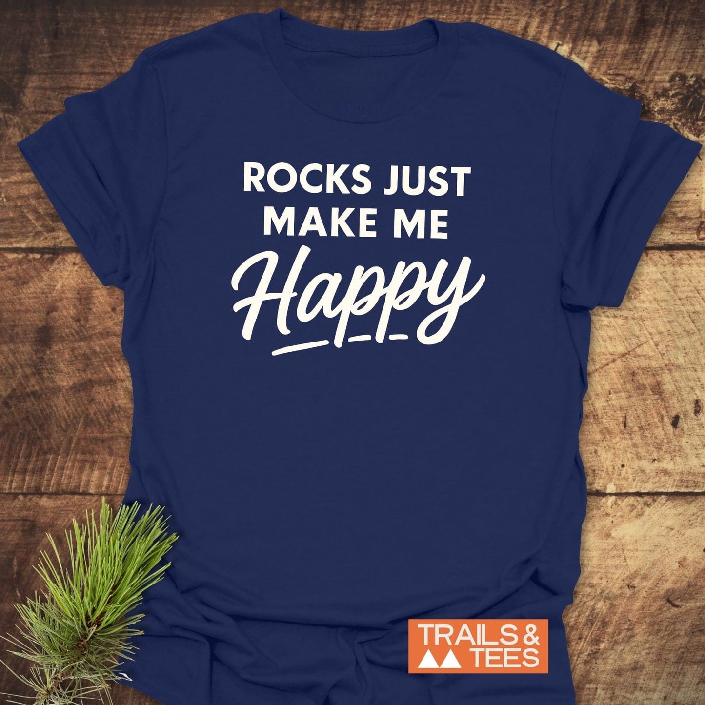 Navy blue Rocks Make Me Happy T-shirt with white lettering, displayed flat on wood. Features durable, soft ring-spun cotton with the Trails & Tees logo.