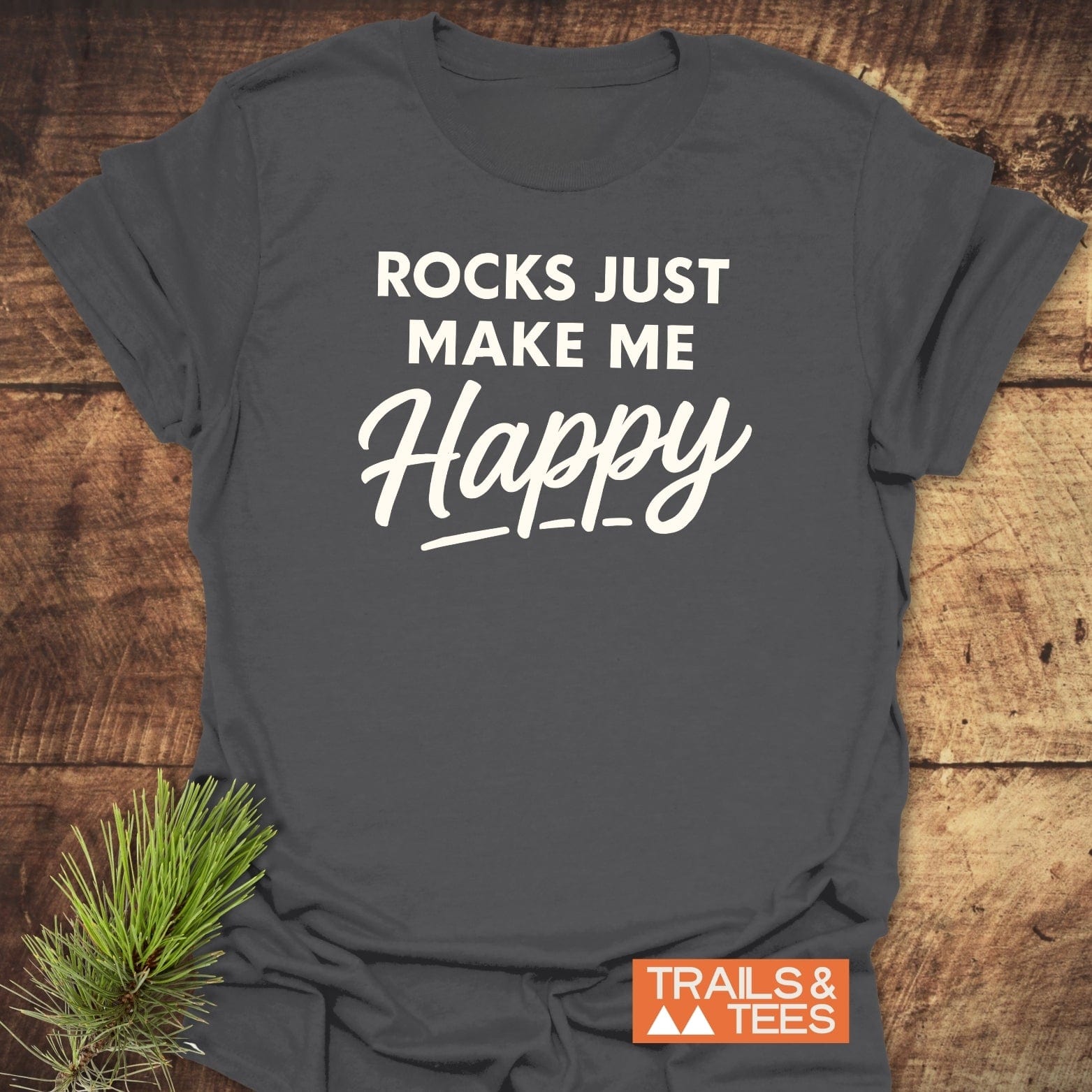 Dark gray Rocks Make Me Happy T-shirt on wood, featuring bold and cursive white print, styled with a pine sprig and Trails & Tees logo.