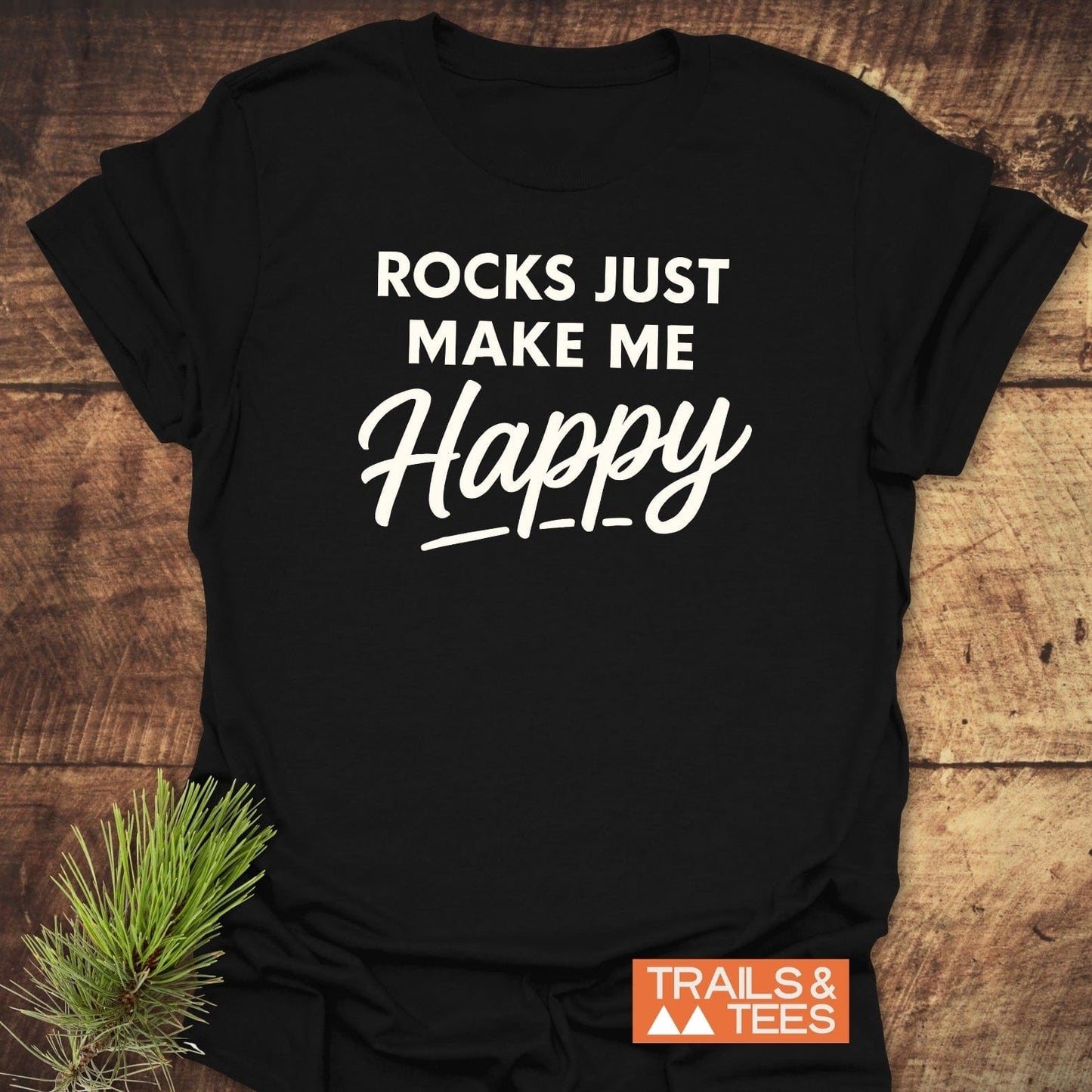Black T-shirt with ROCKS JUST MAKE ME Happy slogan, featuring durable white print. Includes TRAILS & TEES label, pine sprig accent, showcasing outdoor-inspired design.
