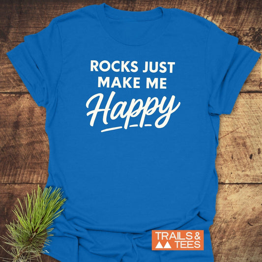 Rocks Make Me Happy T-Shirt on wooden surface, featuring white text, Trails & Tees logo, and pine sprig. Soft, durable, outdoor-inspired design.