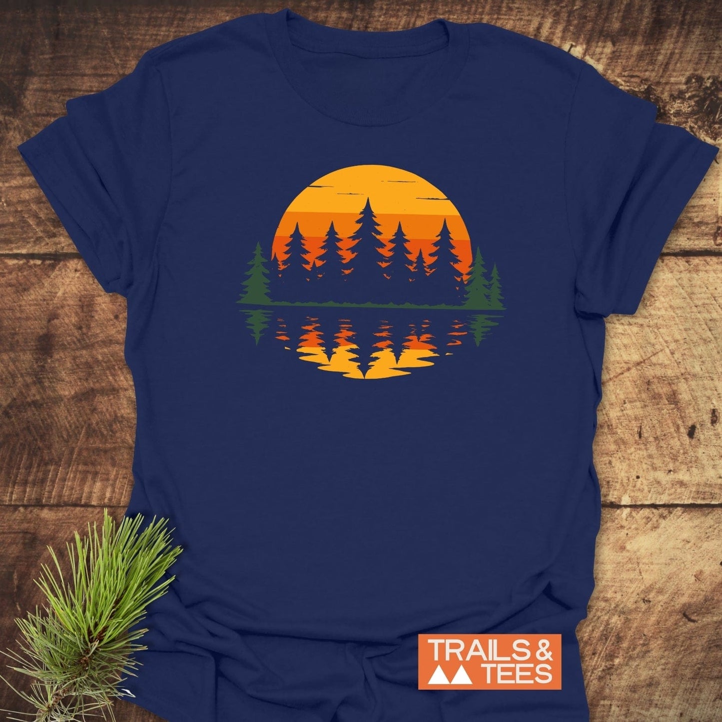 Retro Sunset Pines T-Shirt on a wood surface, features a sunset and evergreen design. Crafted from soft cotton, ideal for outdoor enthusiasts. Includes TRAILS & TEES logo.