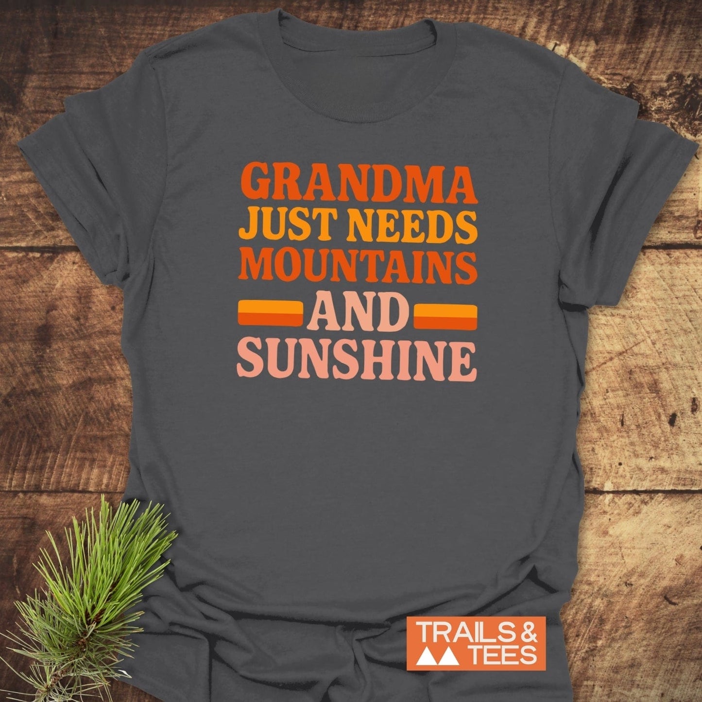 Retro 80s Grandma Mountains T-Shirt on wooden surface with pine sprigs; bold lettering, small orange TRAILS & TEES logo; designed for comfort and outdoor adventures.