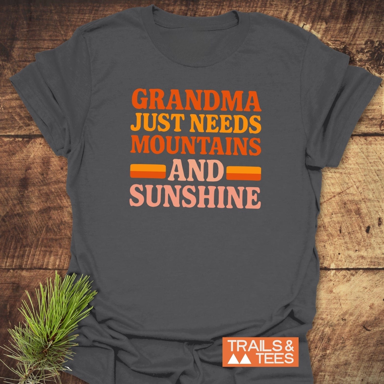 Retro 80s Grandma Mountains T-Shirt on wooden surface with pine sprigs; bold lettering, small orange TRAILS & TEES logo; designed for comfort and outdoor adventures.