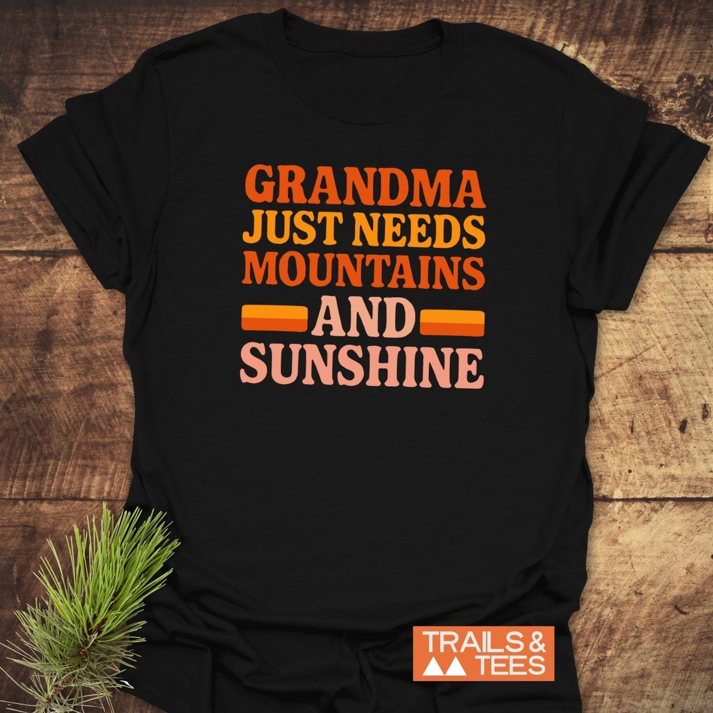 Retro 80s Grandma Mountains T-Shirt on wooden surface, featuring multicolor text graphic. Trails & Tees brand logo visible, with a pine sprig beside the shirt.