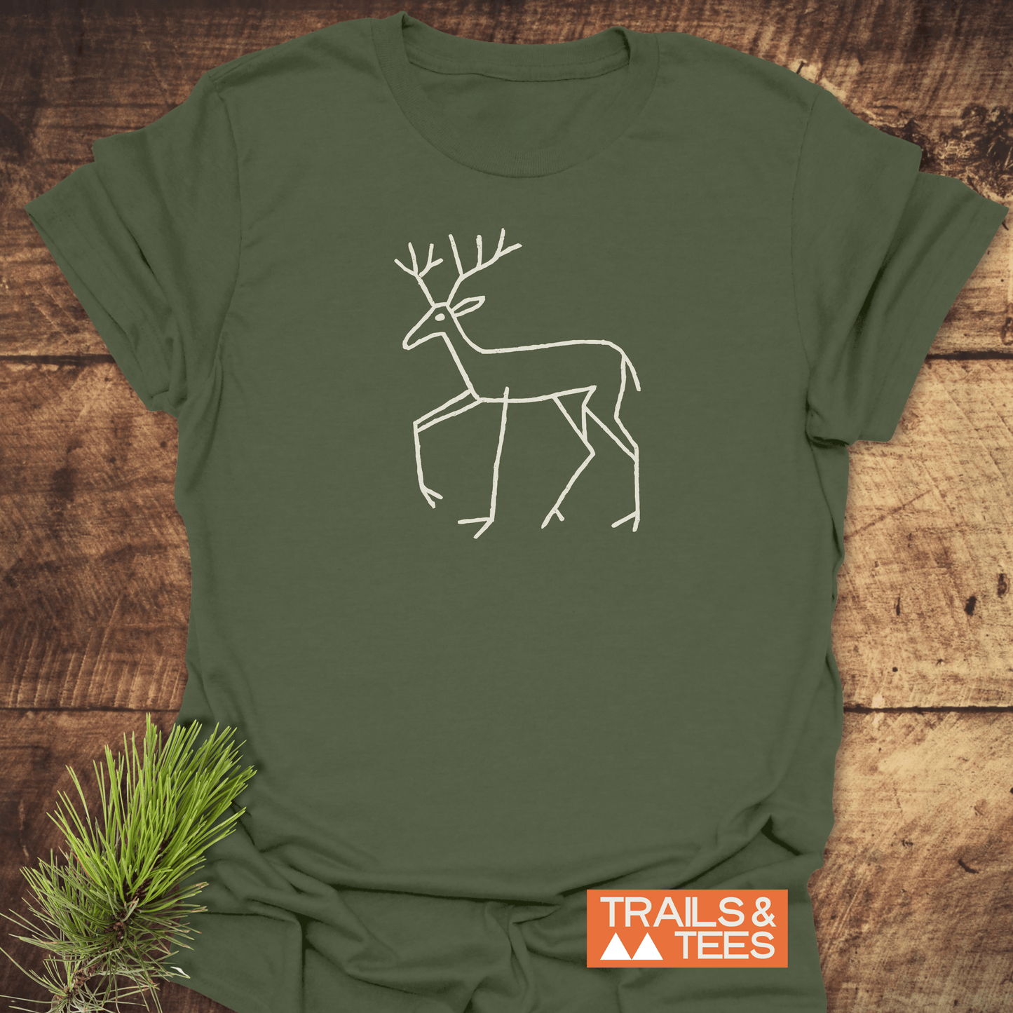 Deer Tribal T-Shirt featuring a white deer line drawing on olive-green fabric, displayed on wood with pine sprig; embodies outdoor-inspired, comfortable adventure wear.