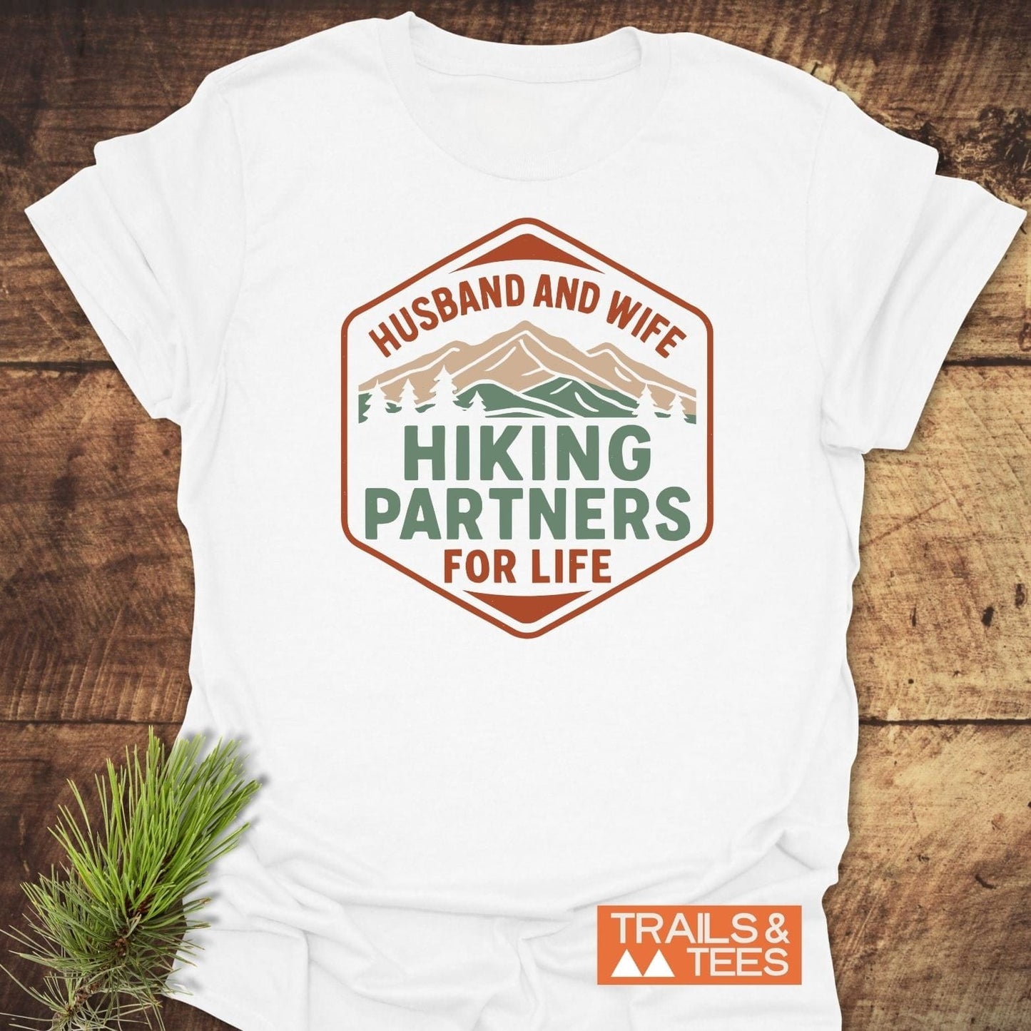 Hiking Partners For Life T-Shirt with earth-tone mountain graphic, soft cotton build, durable prints, and Trails & Tees logo, displayed on a wooden surface.