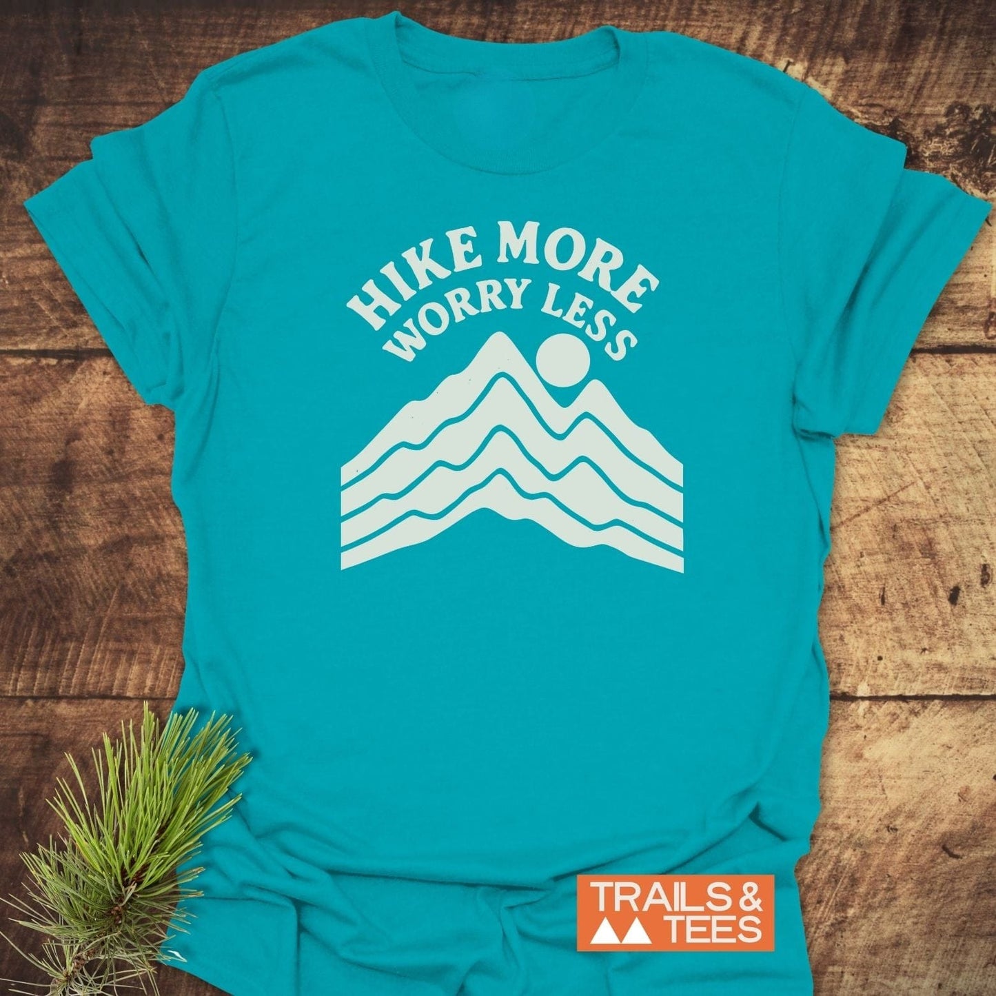 Hike More Worry Less T-Shirt with mountain graphic and sun, displayed flat. Soft cotton, pre-shrunk for fit, logo visible, evergreen sprig accent nearby.