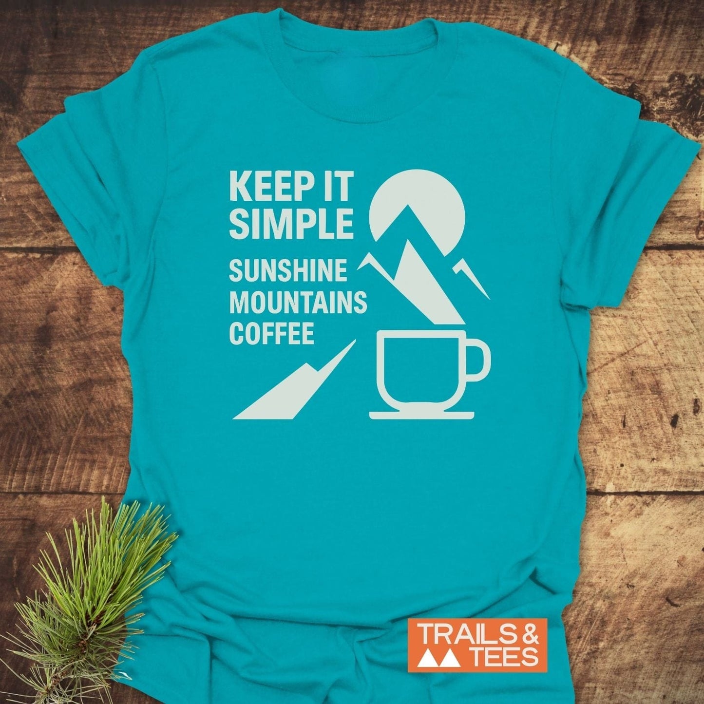 Keep It Simple Mountain T-Shirt featuring white text and icons of sun, mountains, and coffee cup, laid on wood, beside pine sprig.