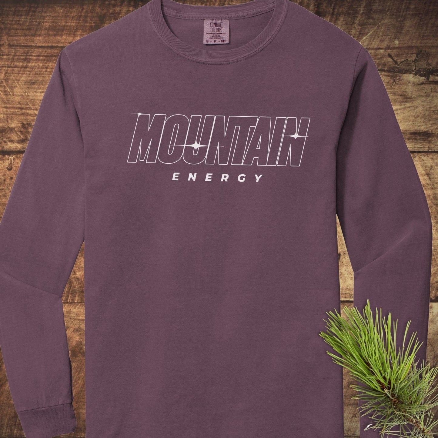 Mountain Energy Comfort Colors Longsleeve Tee, maroon-purple, laid on wood, features bold MOUNTAIN graphic, made of 100% ring-spun cotton for softness and comfort.